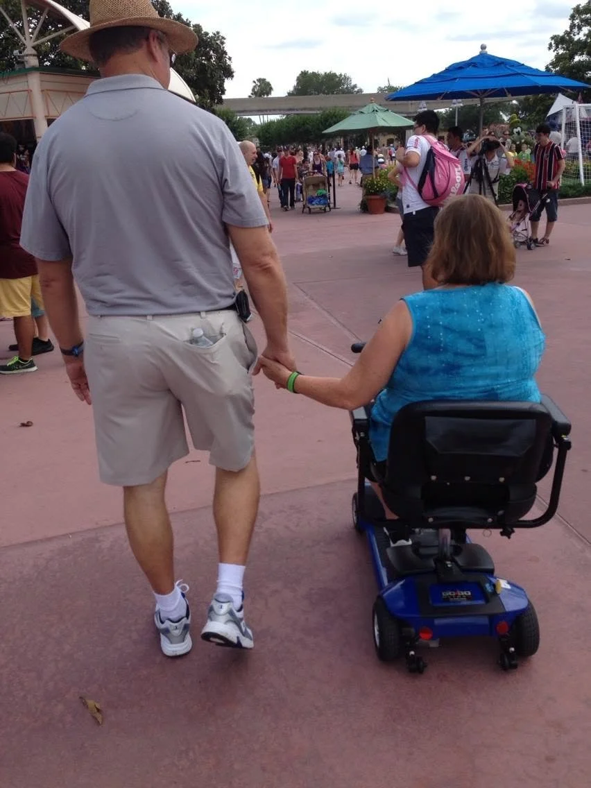 An elderly woman in a blue sleeveless top sits in a mobility scooter, holding hands with a man wearing a gray shirt, beige shorts, and a straw hat at a busy outdoor park or event.