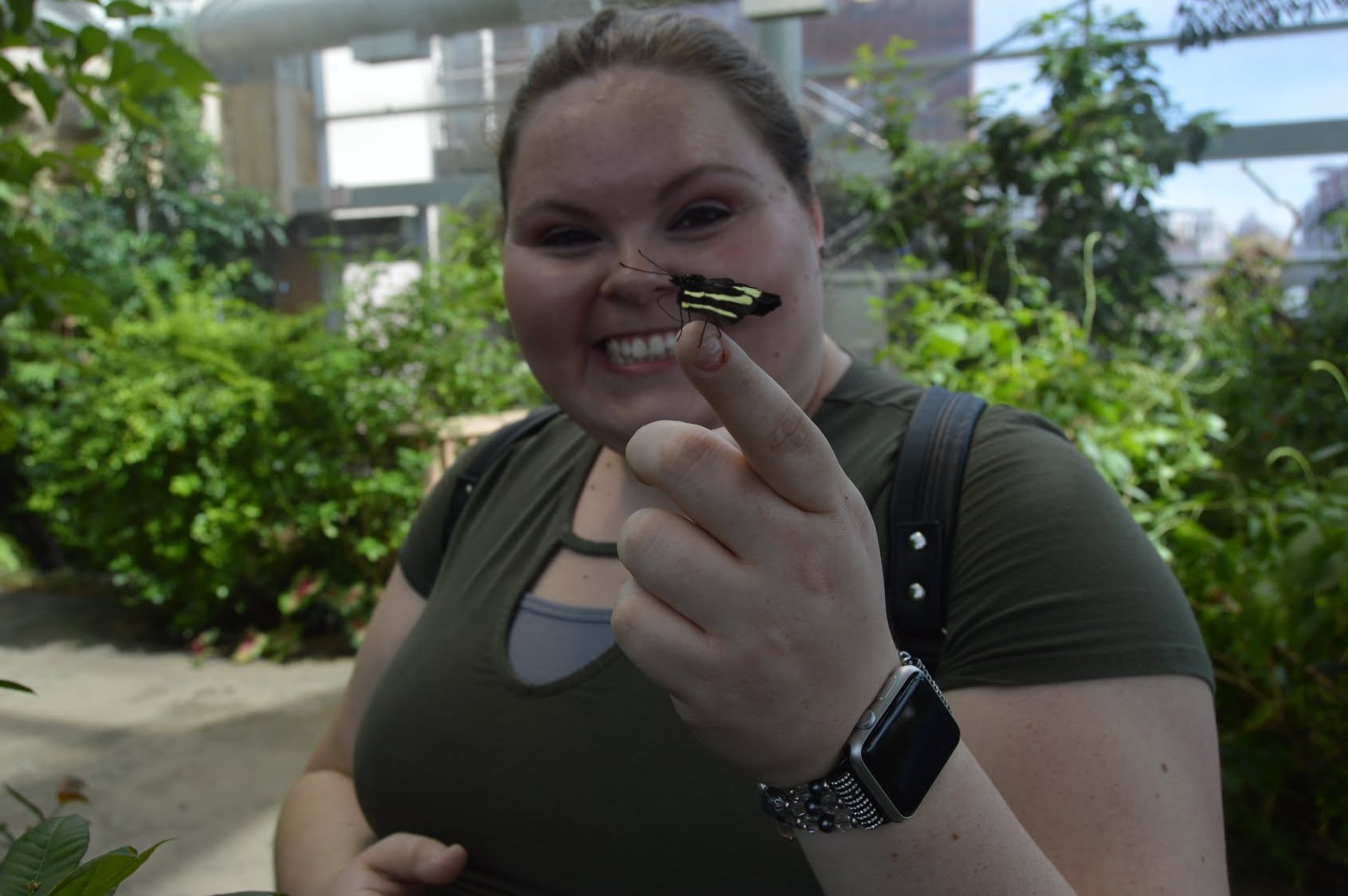 A smiling woman holding a black and yellow striped moth on her finger in a green indoor garden.