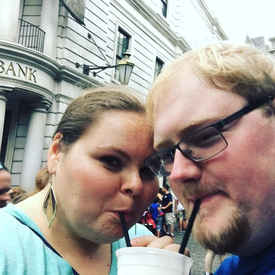Two people drinking beverages through straws in a crowded urban setting with a building that has a sign reading 'BANK' in the background.