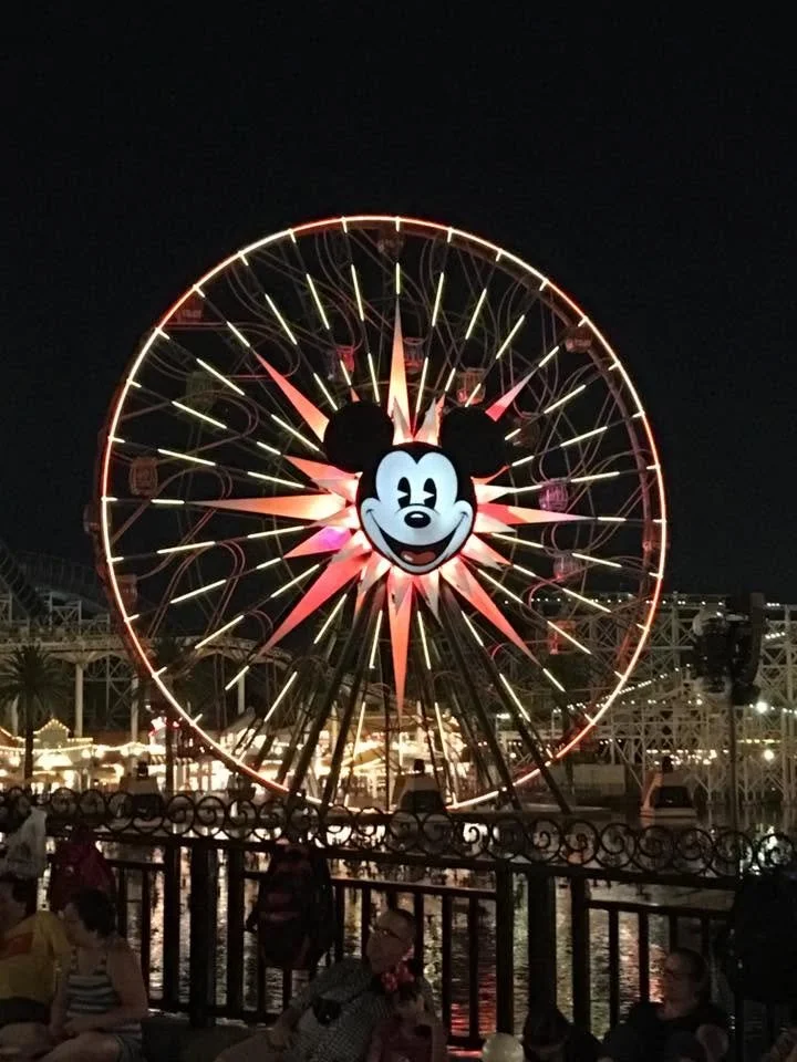 A brightly lit Ferris wheel with a Mickey Mouse face in the center, at night at an amusement park.