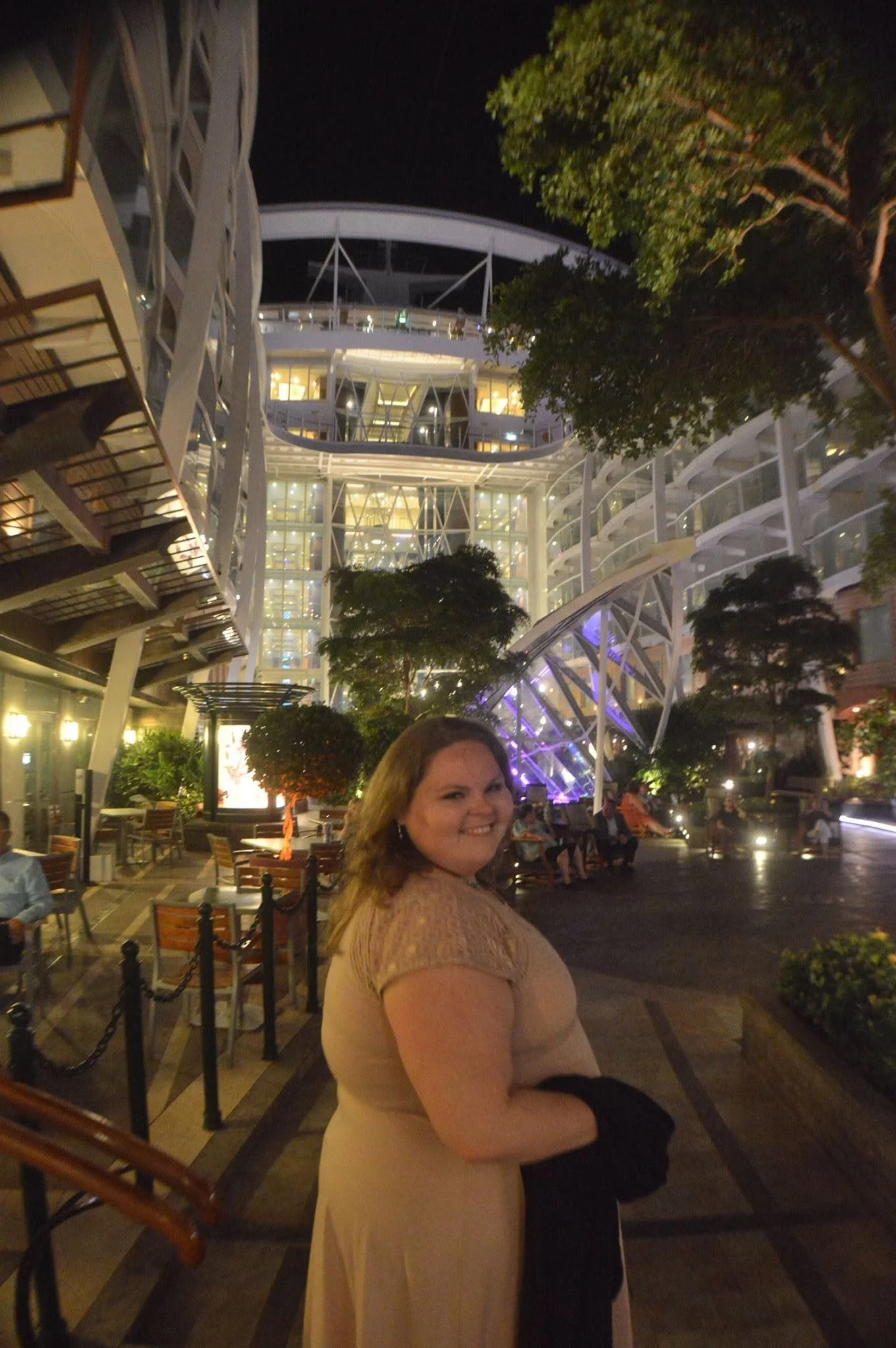 A woman with long hair, wearing a beige dress and black gloves, smiling and posing in an outdoor seating area of a modern building at night with illuminated interiors and lush trees.