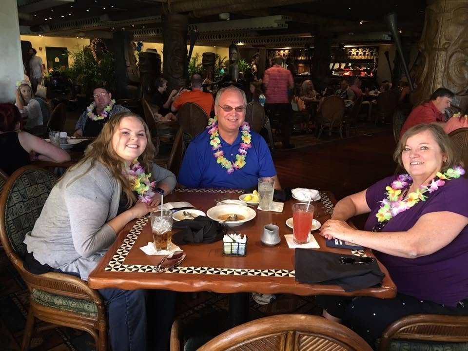 Three smiling people sit at a dinner table in a tropical-themed restaurant, wearing colorful leis and enjoying drinks.