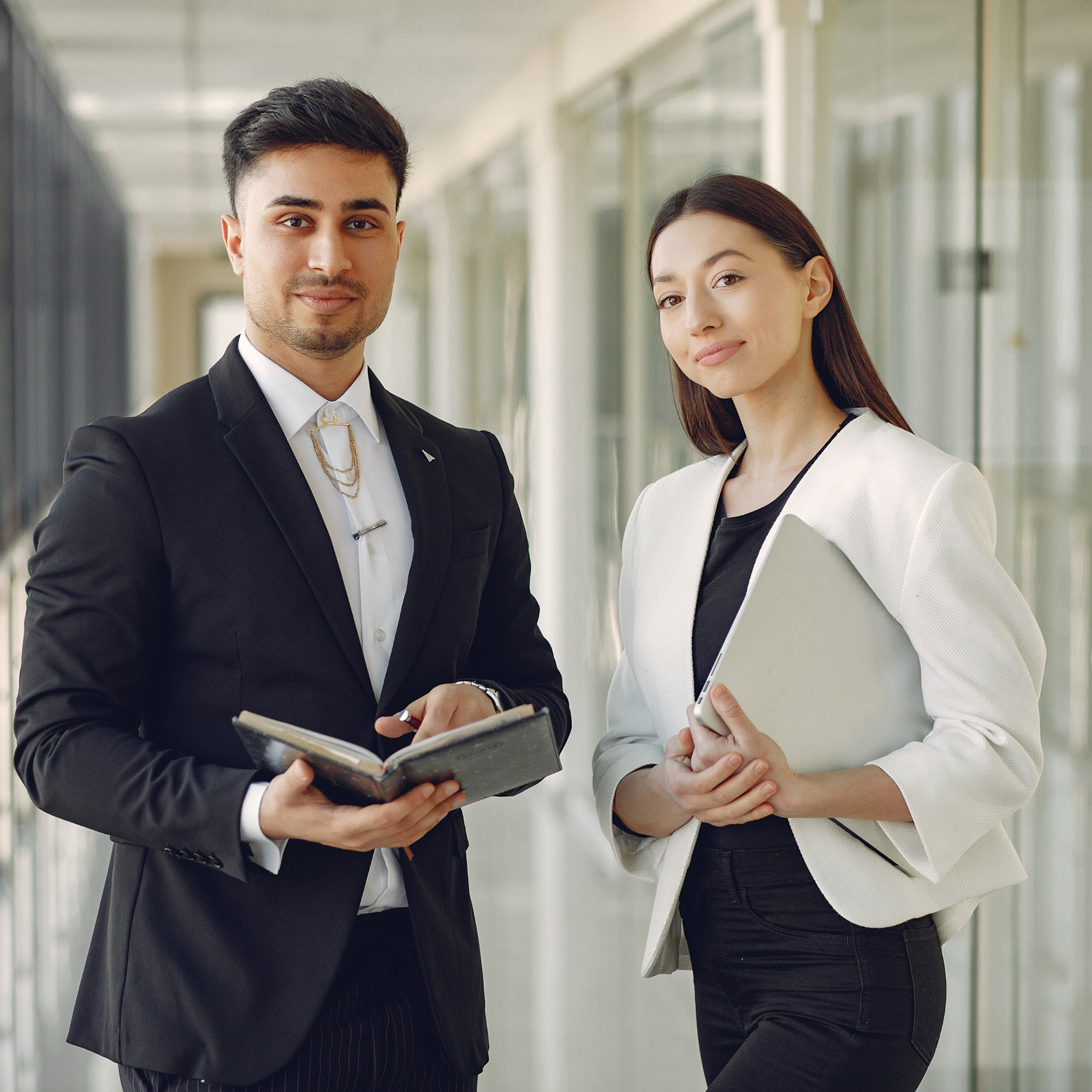A man and a woman standing indoors, dressed in business attire, looking at a notebook or tablet together.