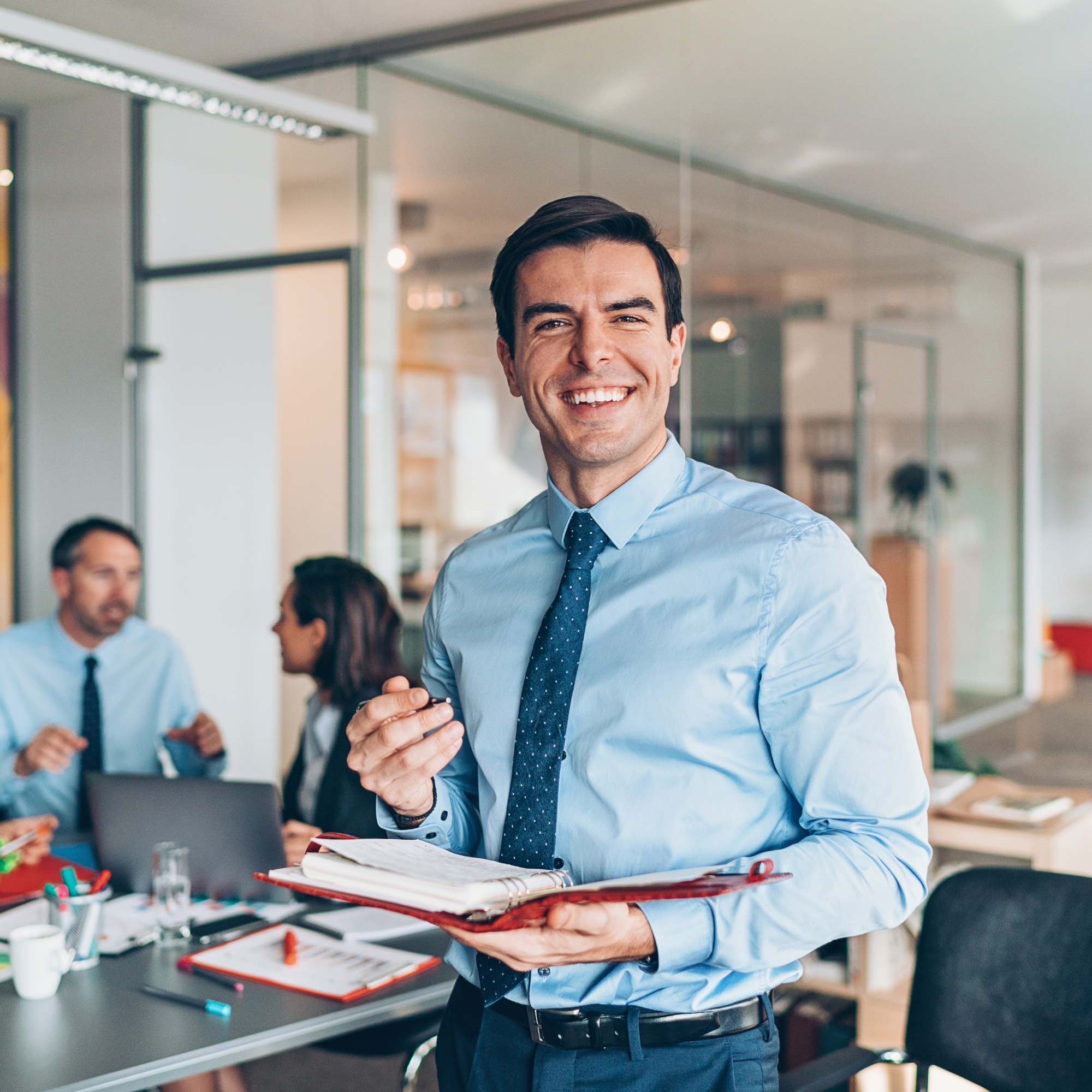 A man in a light blue dress shirt and dark pants is smiling and holding a red folder with papers, standing in an office or conference room with other people in the background.