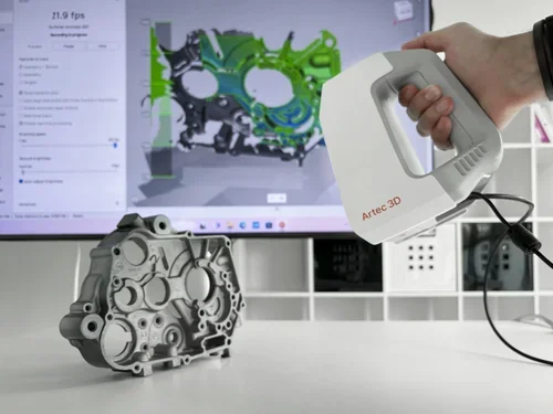 A person holding an Artec 3D scanner over a metal engine part on a white table, with a computer monitor in the background displaying a 3D model of a different engine part.