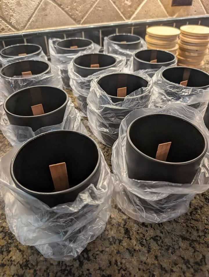 Multiple black glass candle jars wrapped loosely in plastic on a kitchen countertop, with a stack of lids in the background. Ready to hand pour a fresh batch of beautiful botanical candles.
