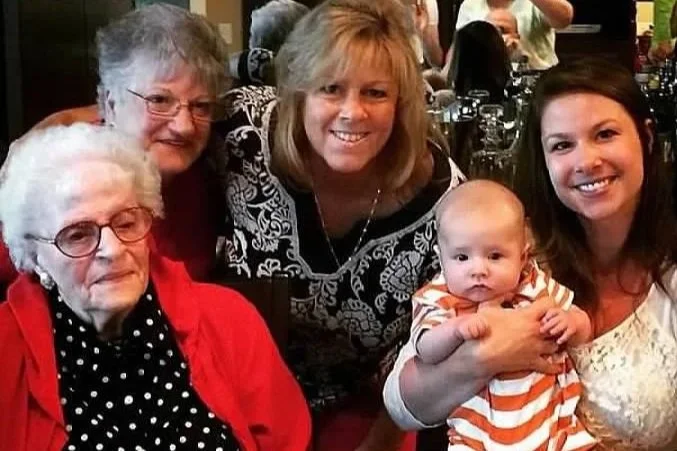 Four generations of women meeting the fifth (a baby) gathered together at a social event, smiling for the camera.