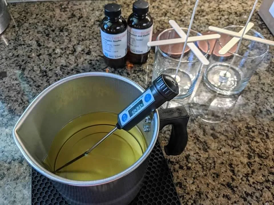 Distillation setup with a temperature probe in a metal pot containing a yellow liquid, two brown bottles labeled 'Klescure,' and three glass jars with copper cups and stirring rods on a granite countertop.