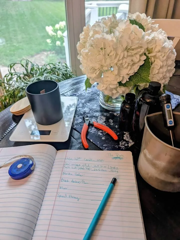 A table with a book, pen, and stationery tools, a vase of white hydrangeas, a black container, and gardening tools, with a window showing a green outdoor scene.