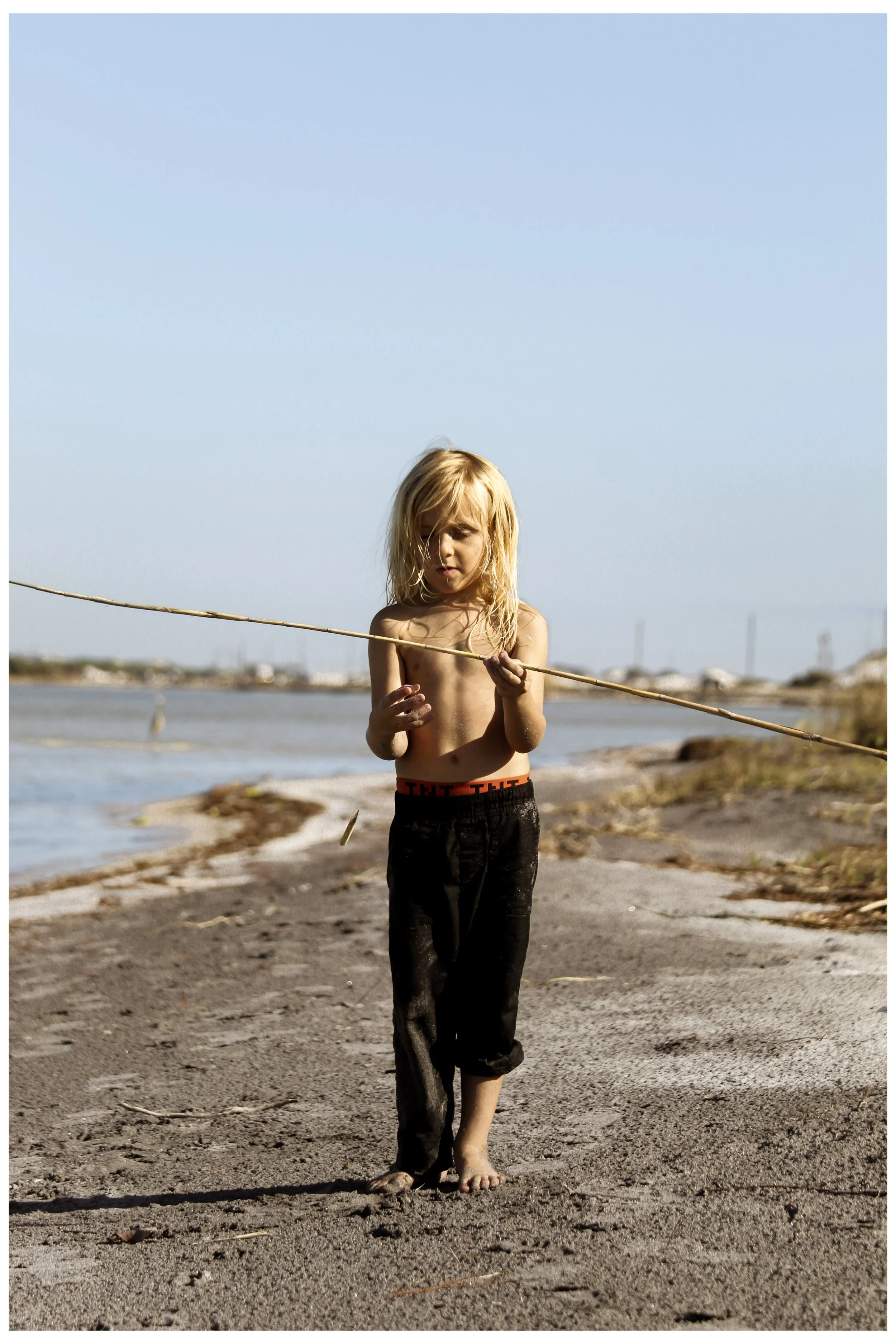 A young boy with long blonde hair, shirtless and wearing black pants, is walking barefoot on a sandy beach. He is holding a long stick and looking down. The background shows water, a clear sky, and distant shoreline with lights or buildings.