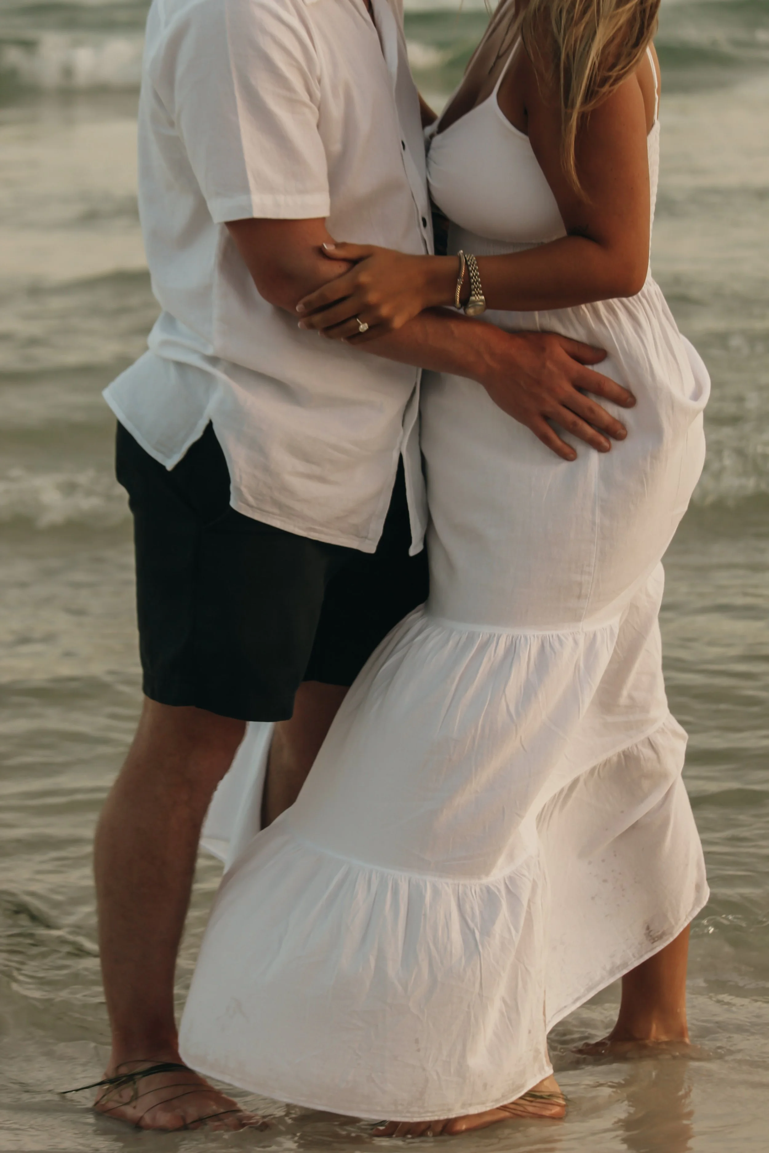 Close-up of a couple standing at the beach, with the woman wearing a white dress and the man wearing a white shirt and black shorts, both with their feet in the water.