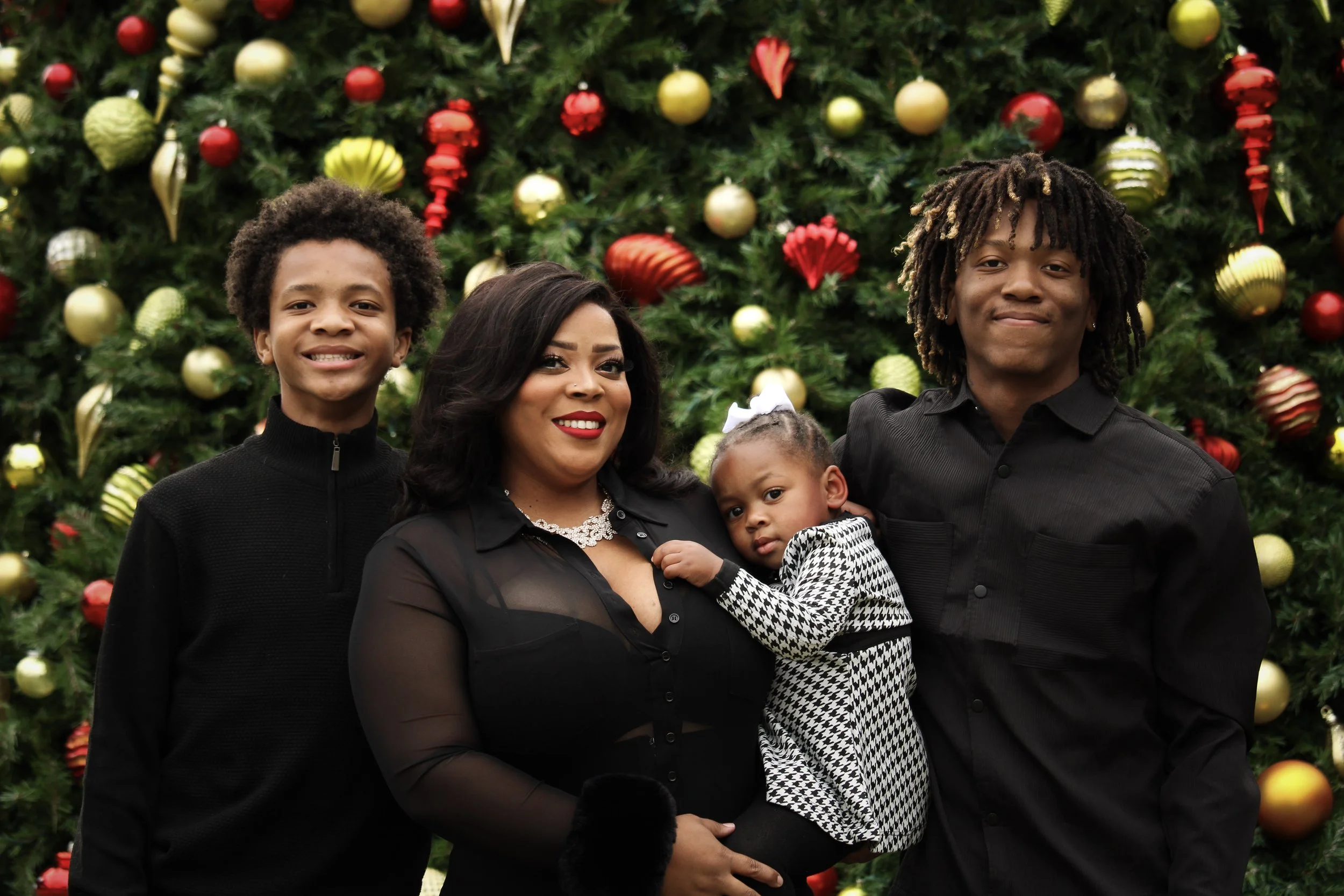 A family of five standing in front of a decorated Christmas tree. The family includes two young men, a woman, and two young children, a girl and a boy. They are smiling and dressed in black.