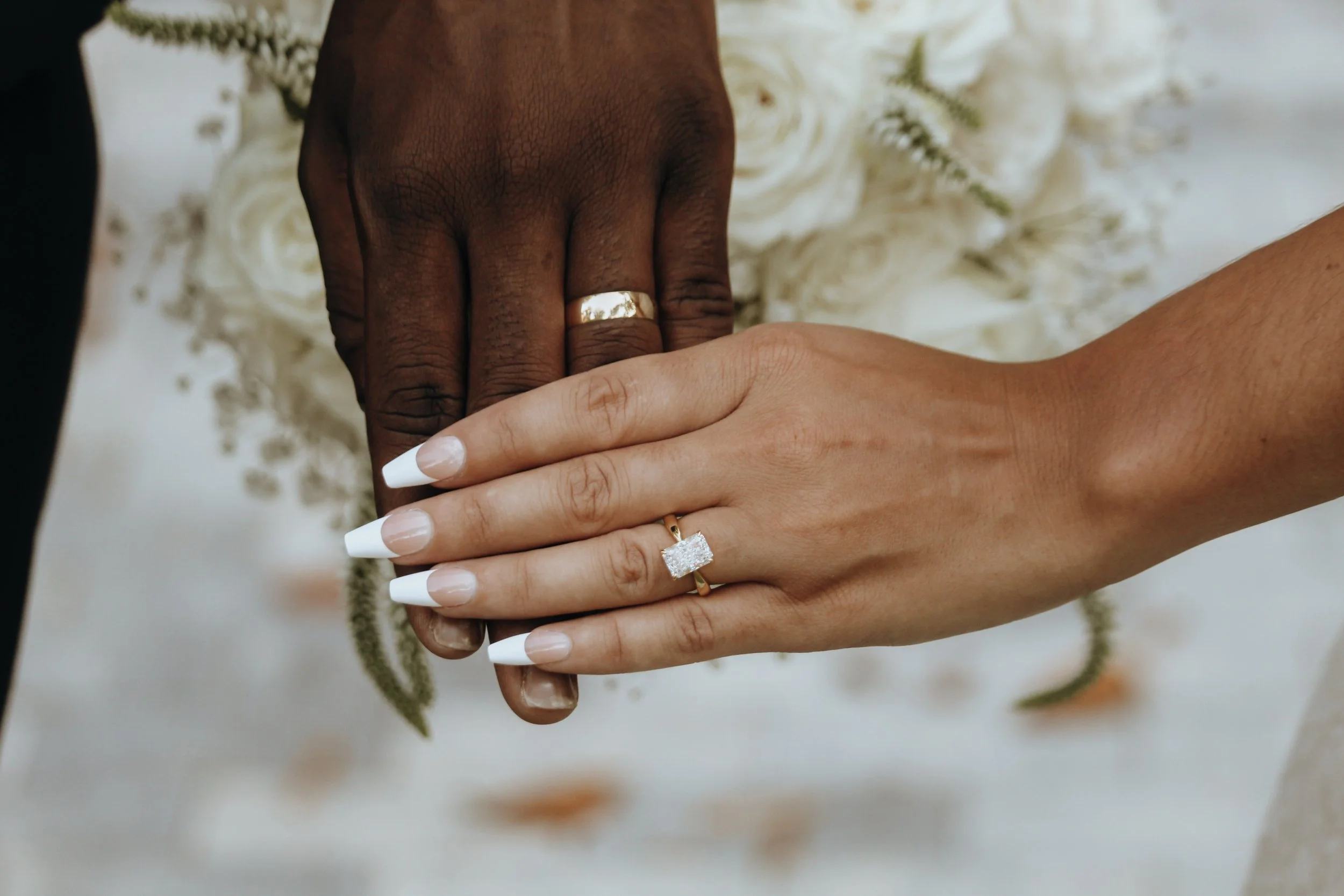 Close-up of two hands, one with dark skin and one with light skin, gently holding each other. The person with dark skin is wearing a gold wedding band on their ring finger. The person with light skin has a large, square-cut diamond engagement ring an
