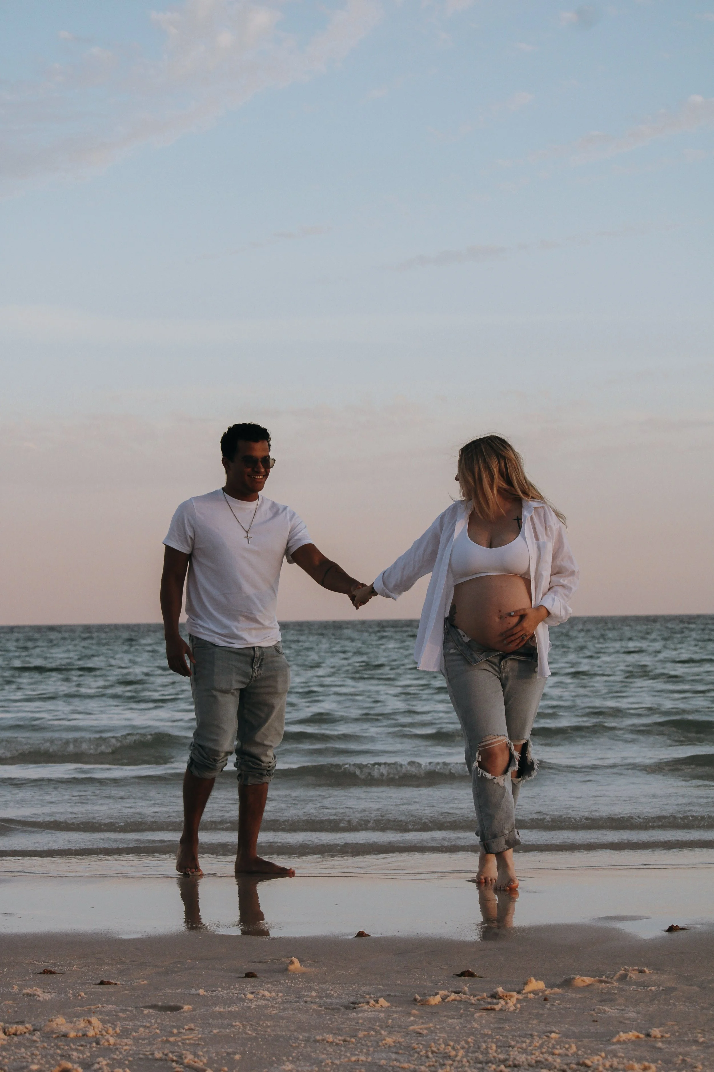 A man and a pregnant woman holding hands on the beach during sunset.