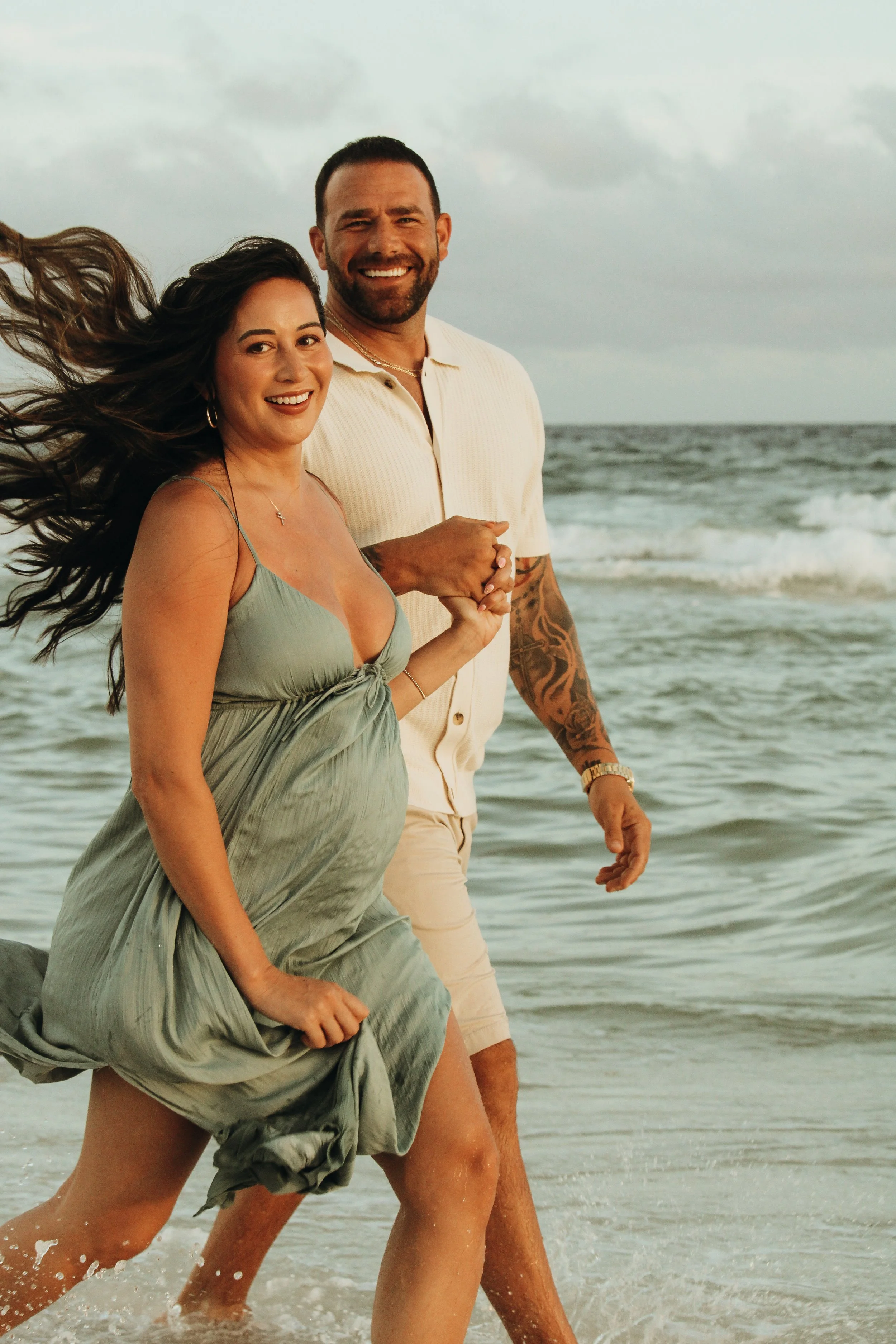 A couple happily walking along the beach near the ocean, with waves in the background and cloudy sky.