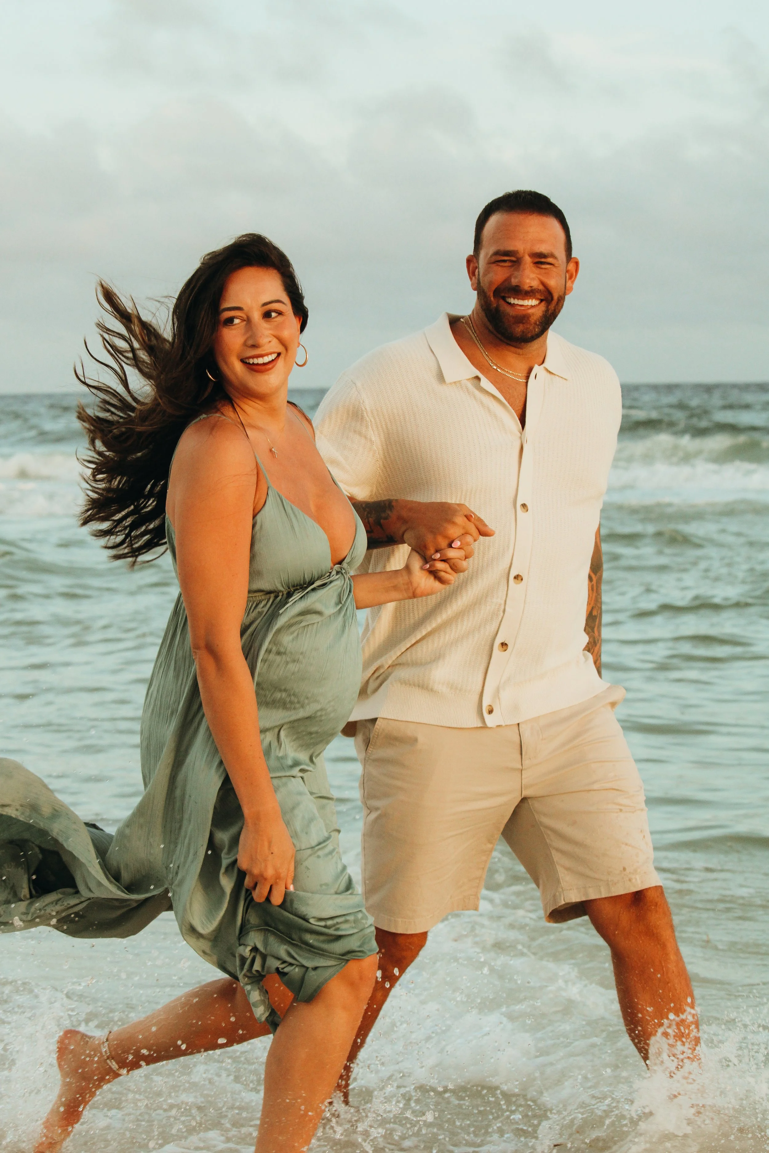 A smiling couple holding hands, walking along the shoreline at the beach, with waves crashing around their legs under cloudy skies.