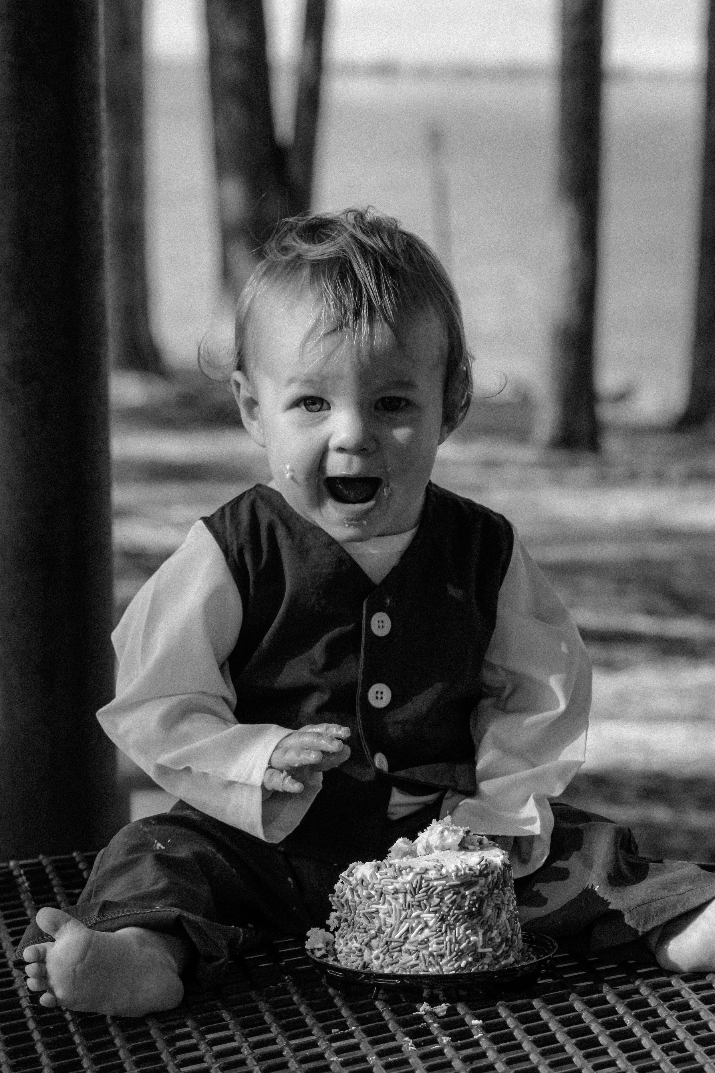A young child with messy hair and food on their face, sitting outdoors and looking at a cake with sprinkles, possibly celebrating a birthday.