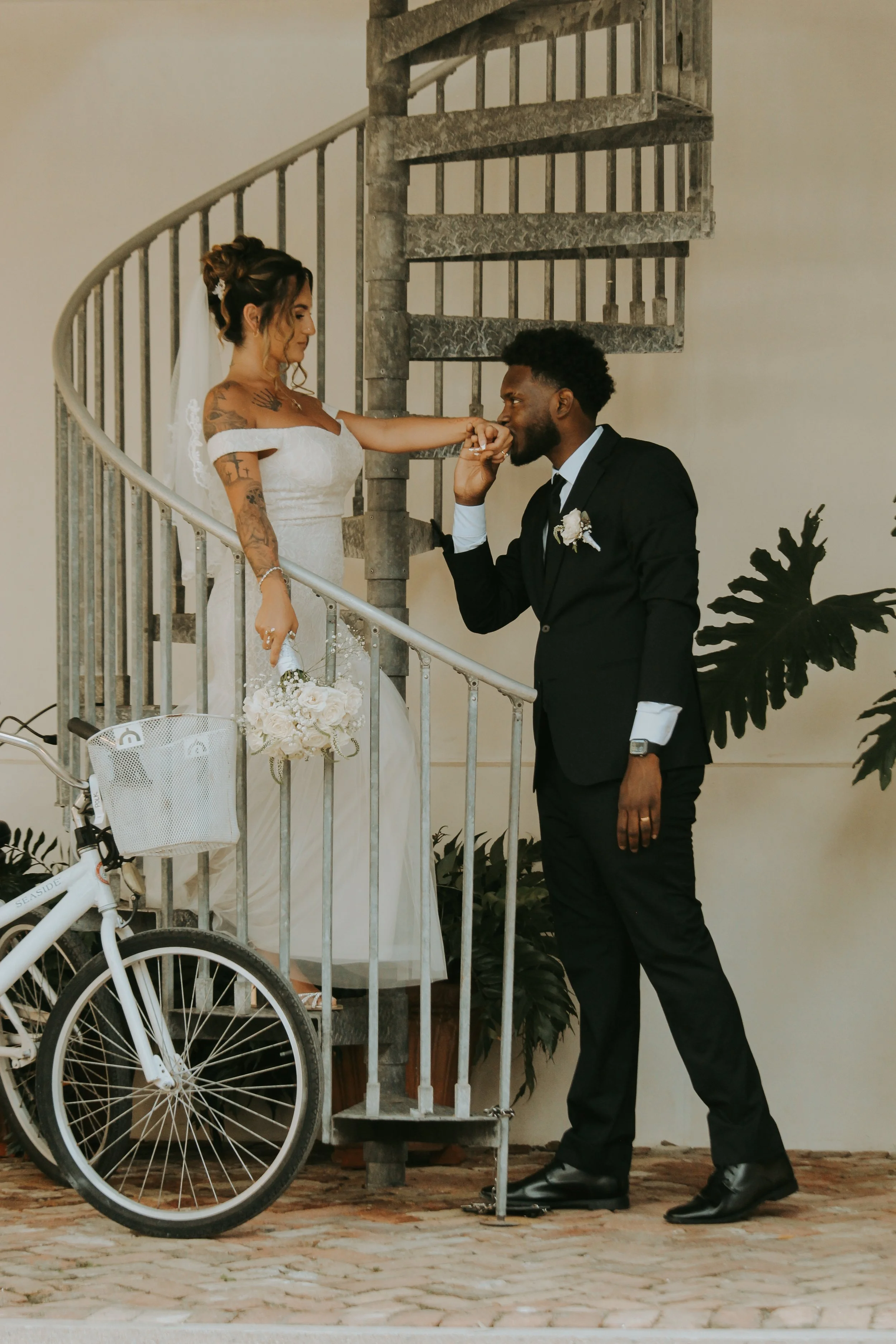 A couple dressed in wedding attire on a spiral staircase. The bride, with tattoos, is touching the groom's hand as he kisses her knuckles. The groom is in a black suit with a boutonniere and is kneeling on one knee. A white bicycle with a basket is v