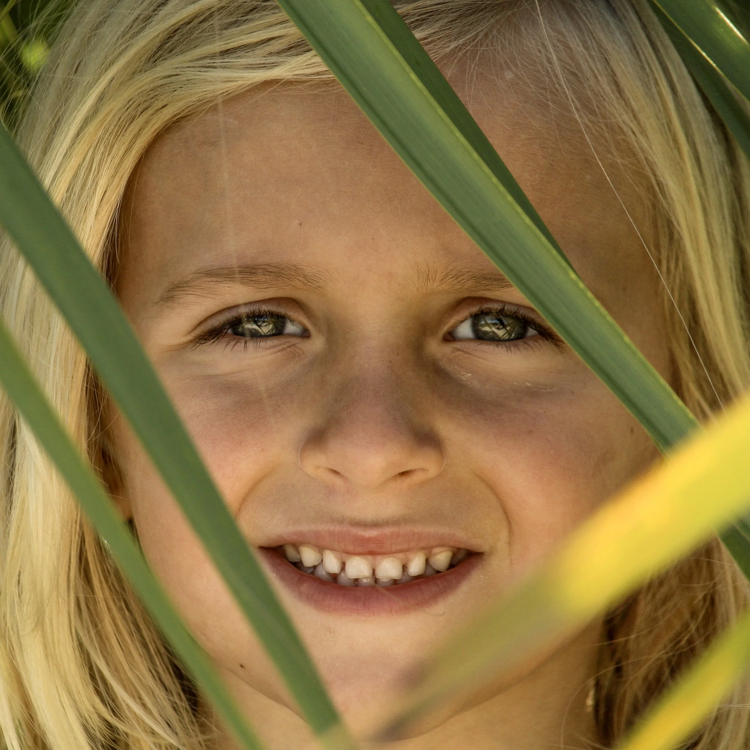 Close-up of a young woman with blonde hair smiling, partially obscured by green leaves.
