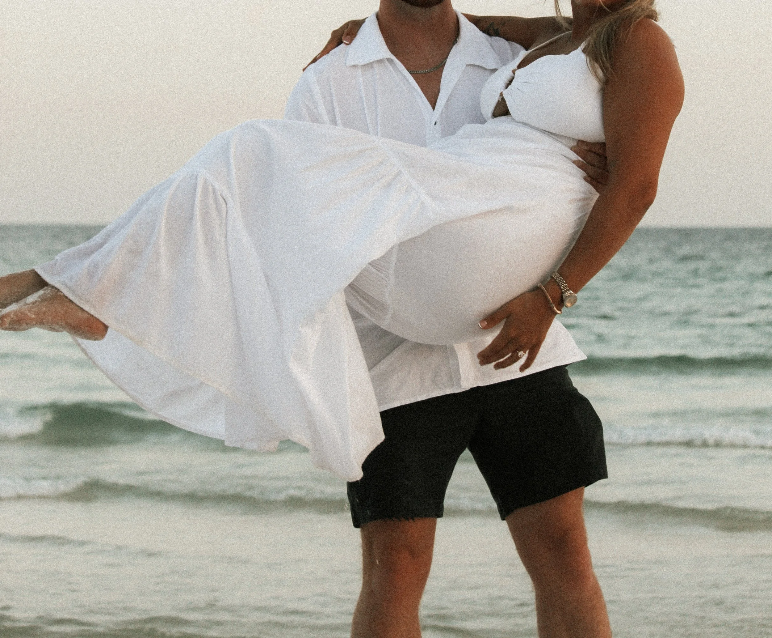 A couple at the beach, with the man carrying the woman, both dressed in white and black summer clothes, with ocean in the background.