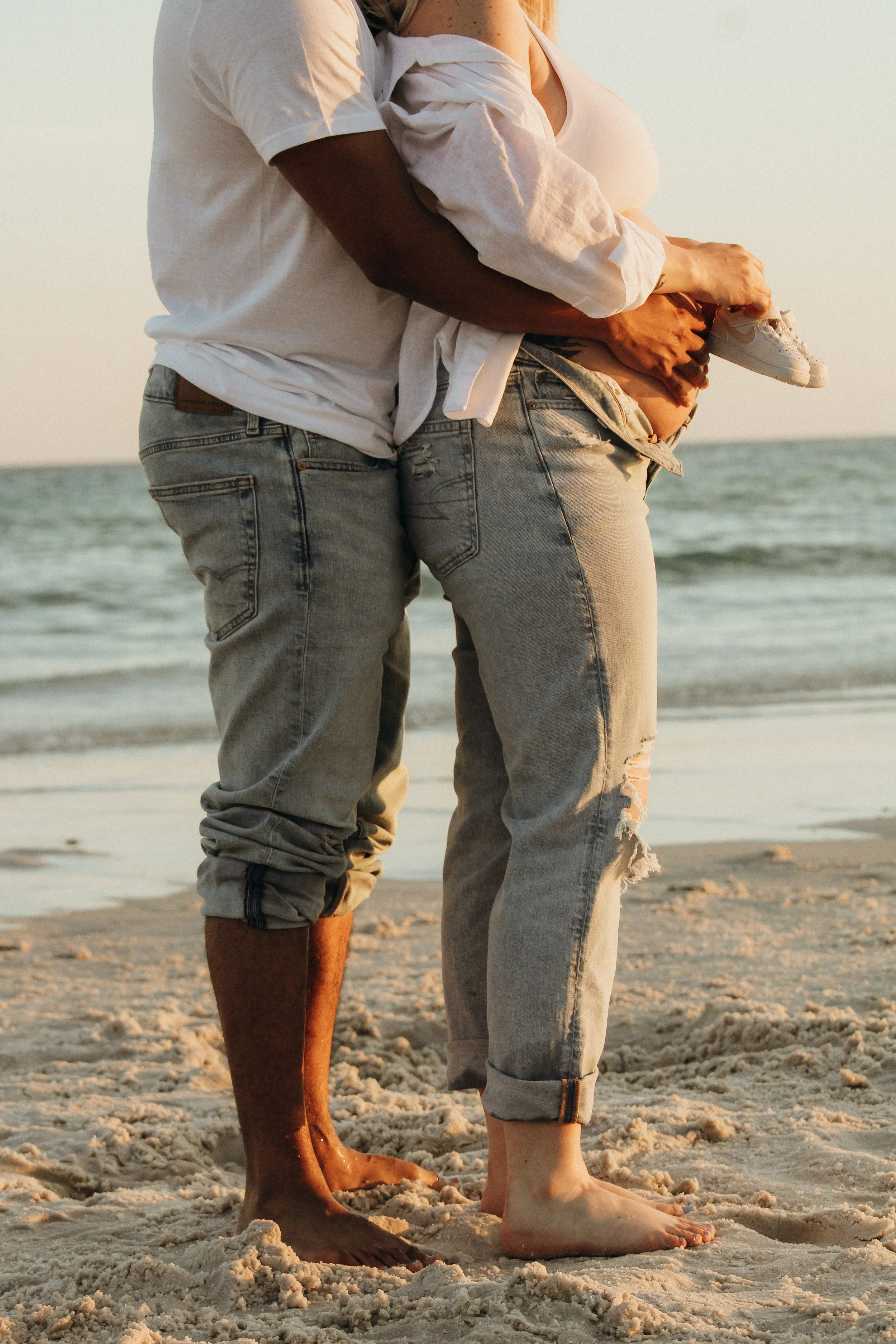 A couple standing on a beach with their feet in the sand, embracing each other as the sun sets over the ocean.