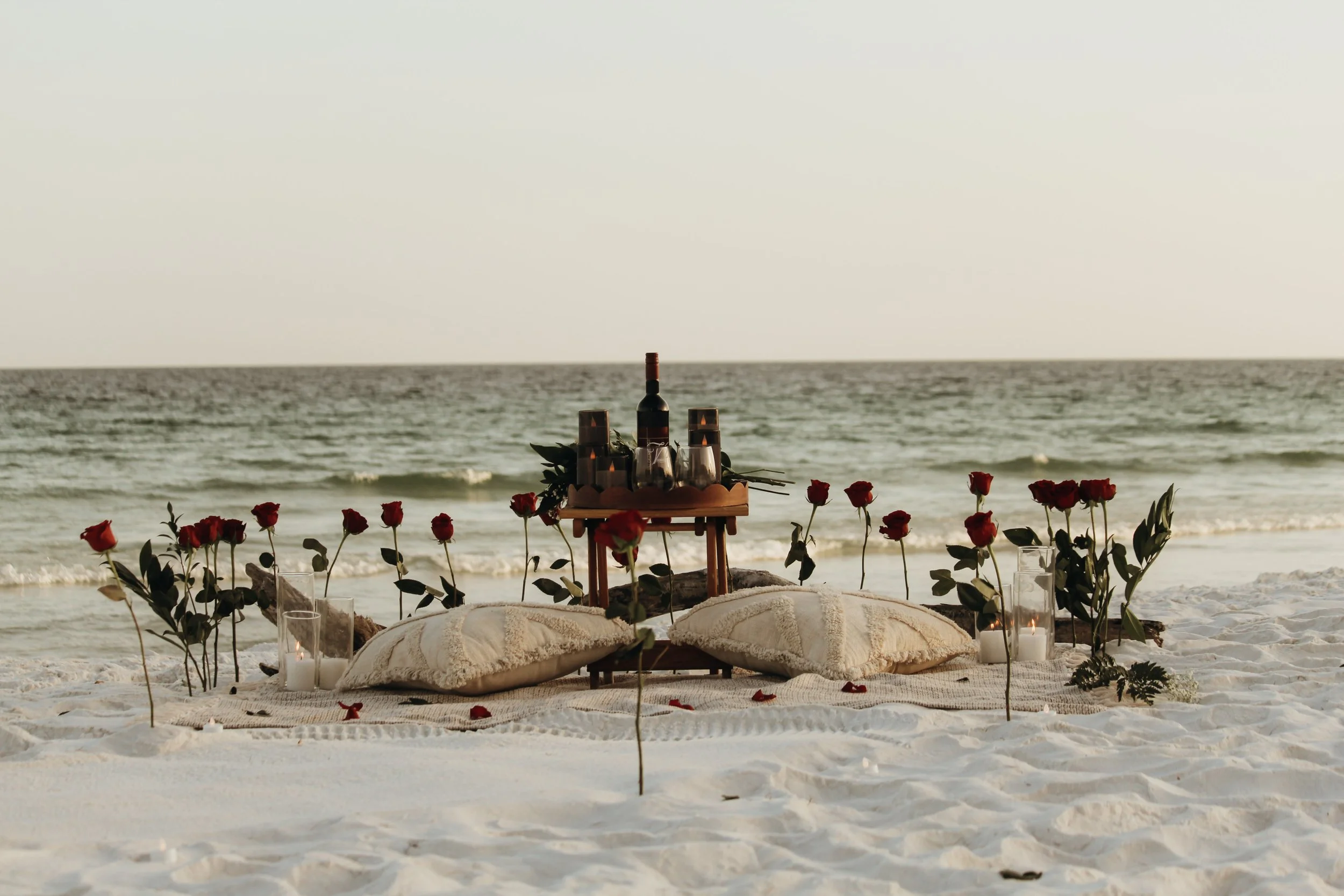 A romantic beach setup with wine bottles, glasses, and roses arranged on a wooden table, surrounded by candles, on sandy beach with the ocean in the background.