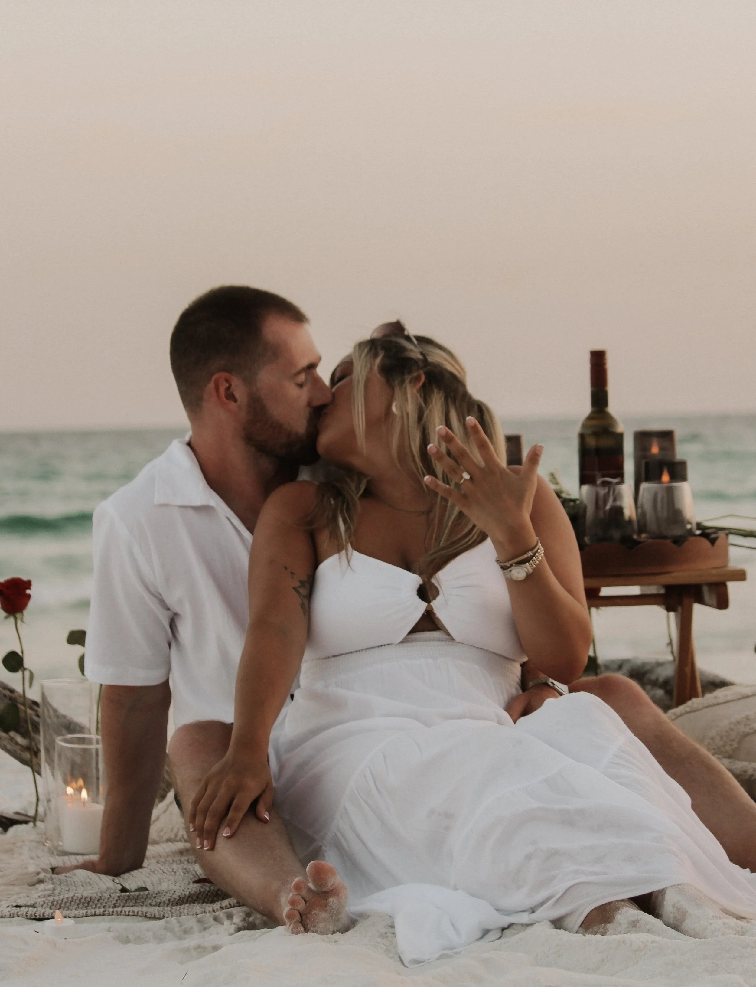 A couple sharing a kiss while sitting on the beach during sunset, with a table of drinks and candles in the background.