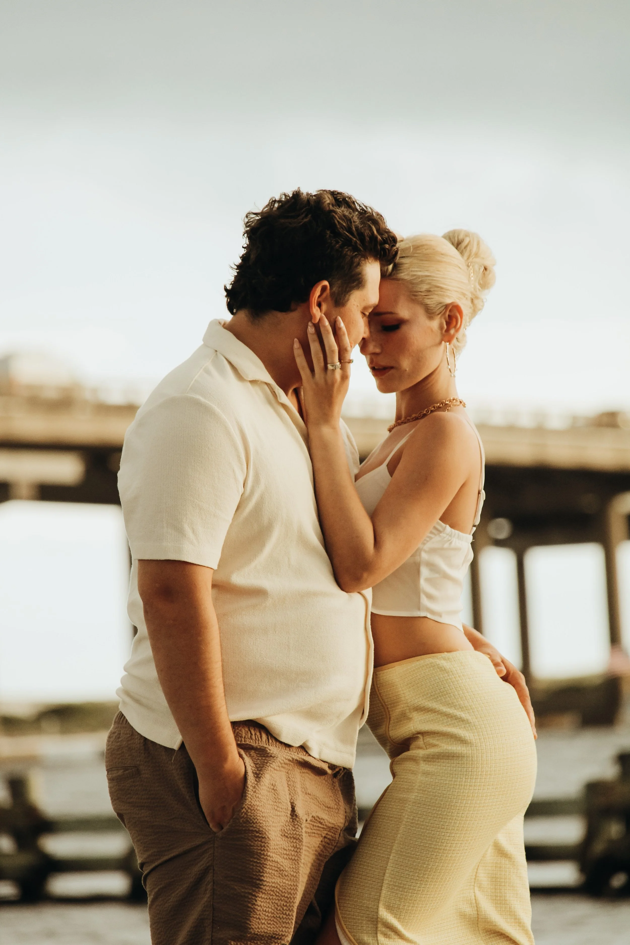 A couple standing close on a beach, with their foreheads and noses touching, and the woman touching the man's face.