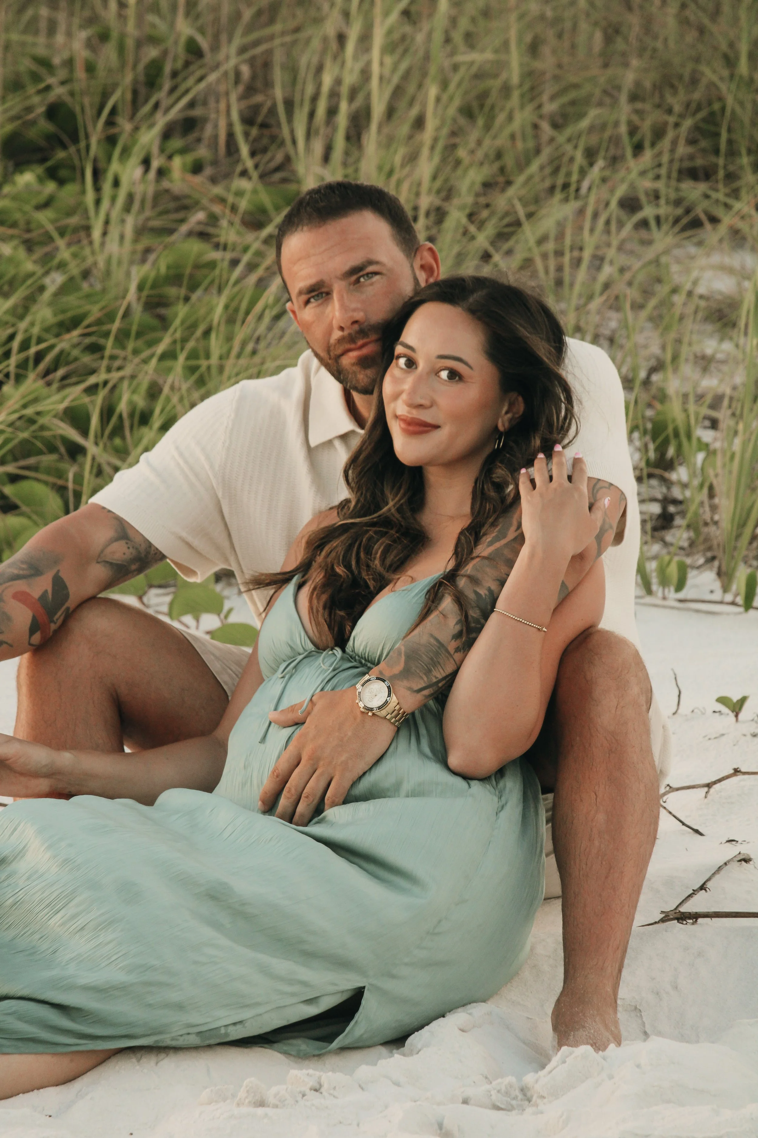 A couple sitting on the sandy beach, with the woman sitting on the man's lap, surrounded by greenery and sand, with relaxed expressions.