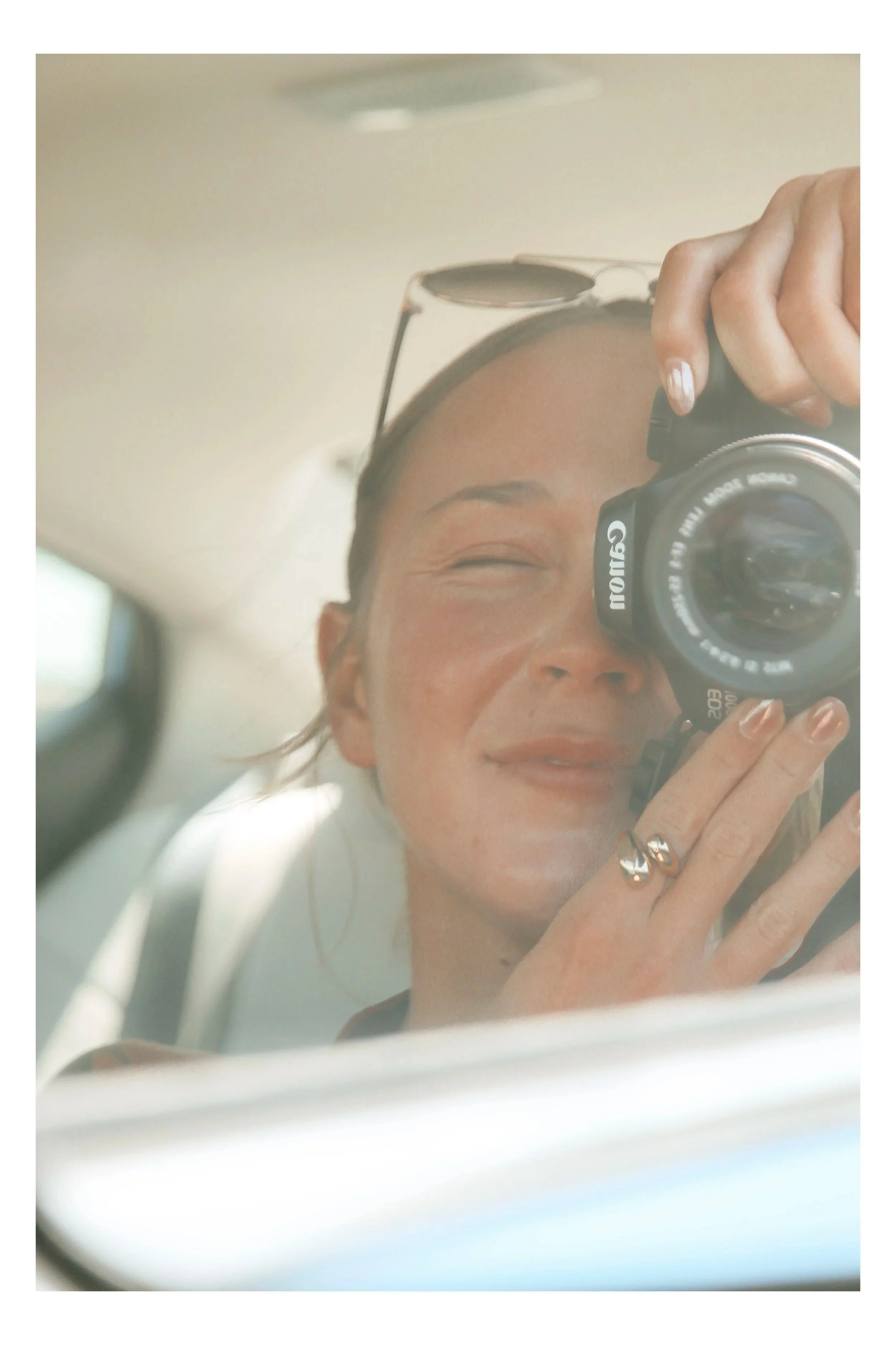 A woman taking a selfie in a mirror with sunglasses on top of her head, smiling with closed eyes, showing curly hair, wearing rings, and holding a camera