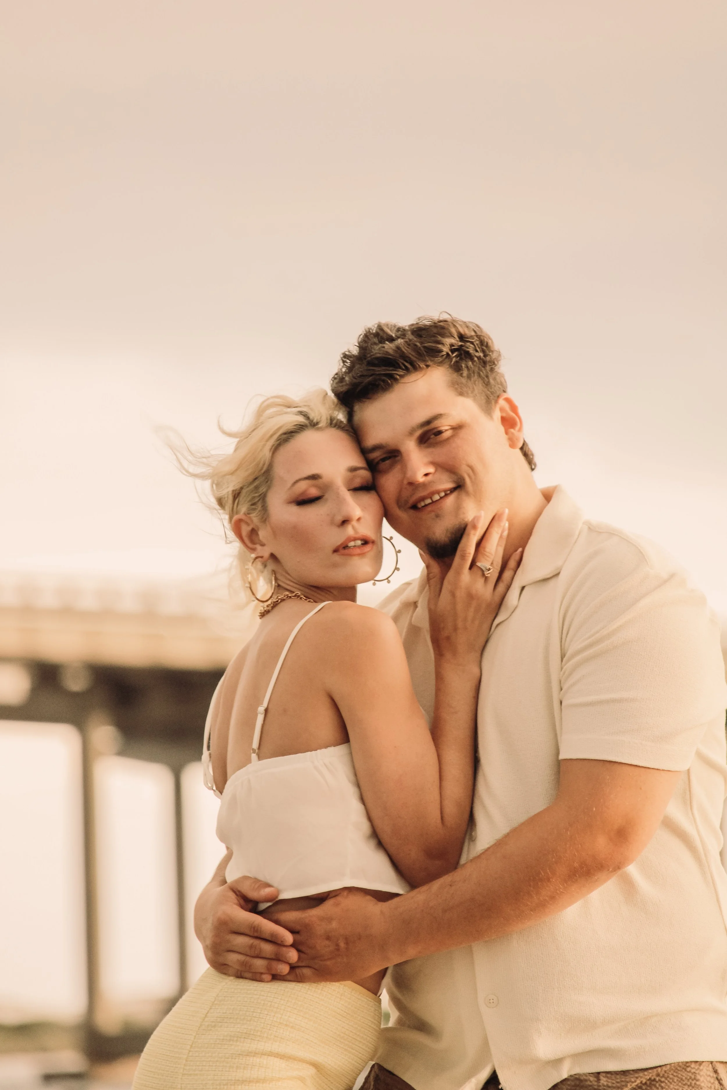 A man and a woman are embracing each other closely in a warm, intimate moment. The woman has blonde curly hair, and the man has short, dark hair. They are smiling gently, with the woman resting her head on the man's forehead. The background is softly