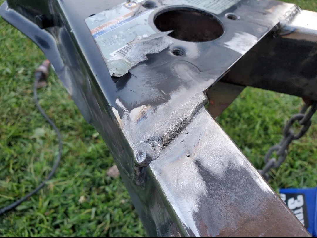 Close-up of a metal welding on a steel frame, outdoor setting with grass. The weld connects two metal pieces, with visible weld beads.