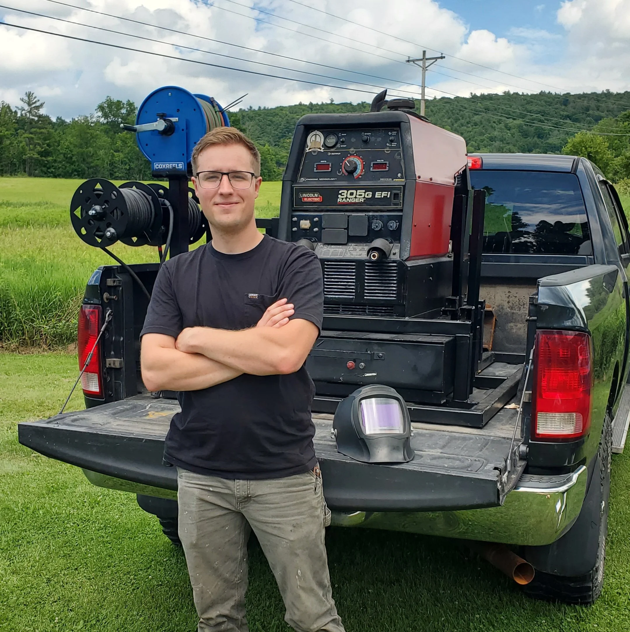 A man standing outdoors with crossed arms next to a pickup truck equipped with welding equipment, in a rural area with green grass, trees, cloudy sky, and power lines in the background.