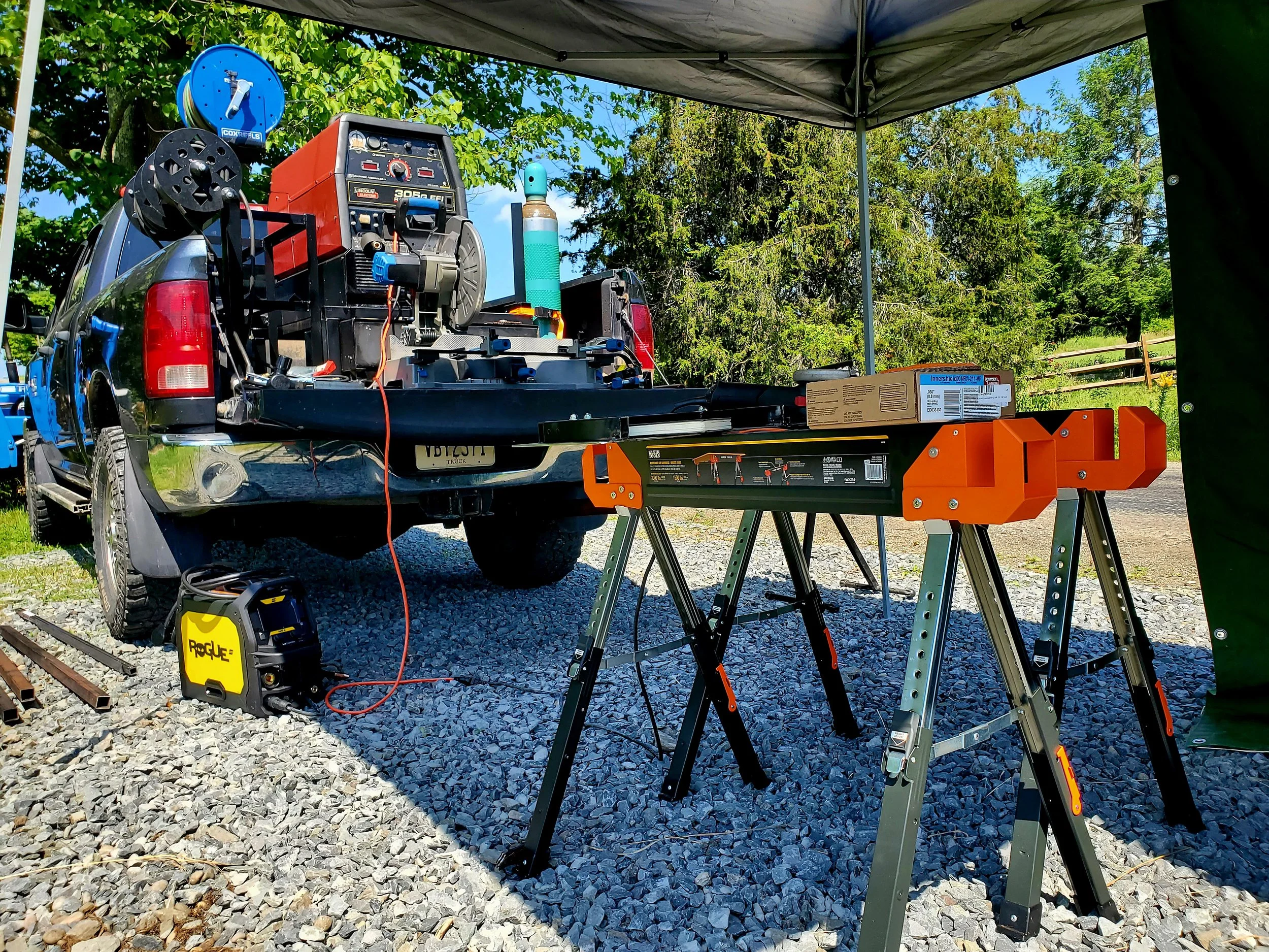 The image shows work setup outdoors under a black canopy, with a truck bed containing tools and equipment, including a welding machine, extension cords, a tank, and a work table. There are trees and a fence in the background, indicating a rural or suburban setting.