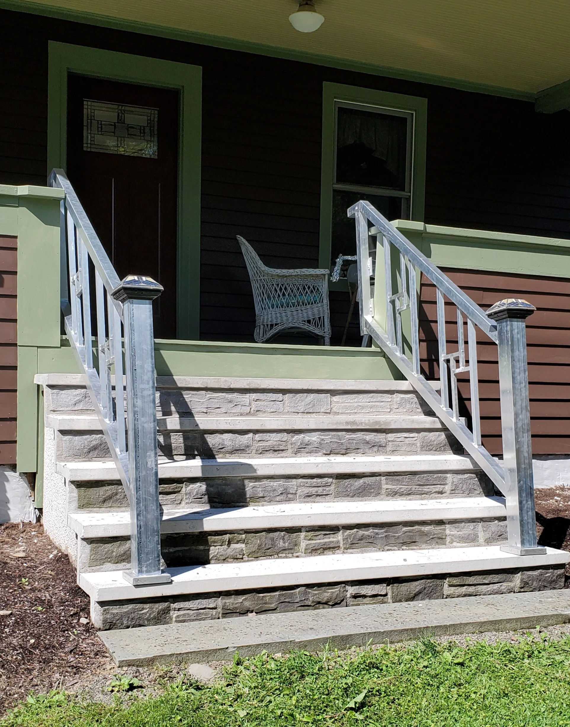 Front porch with concrete stairs, metal railing, a wicker chair, and a green and brown house with a door, window, and a ceiling light.
