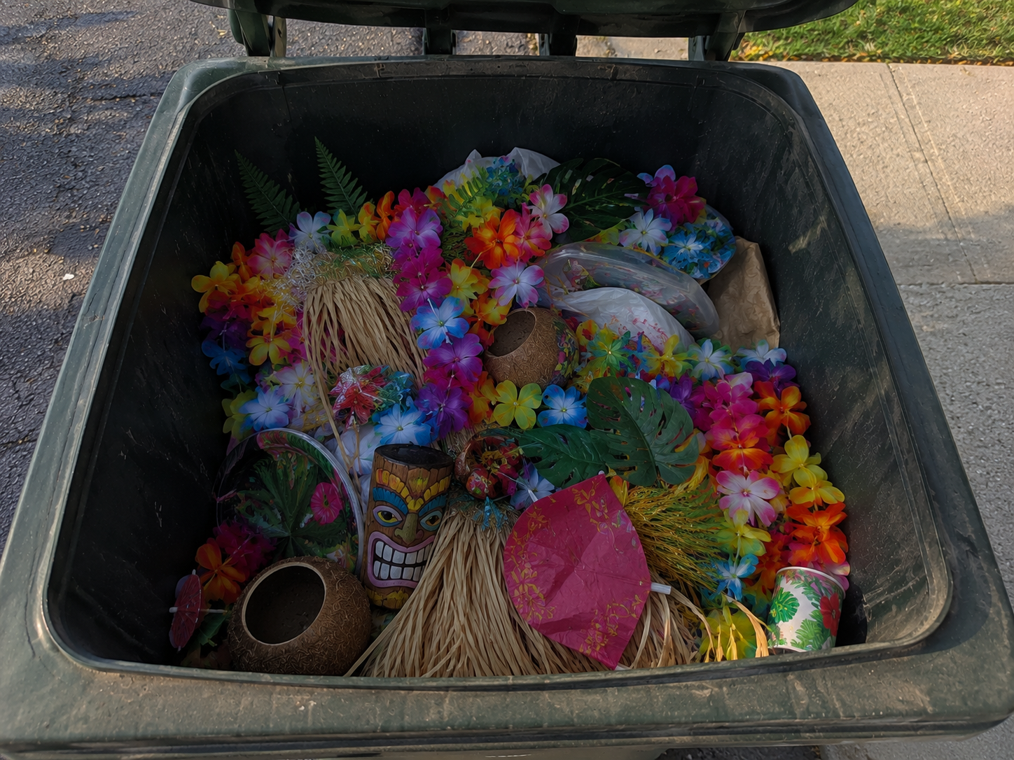 A trash bin filled with colorful Hawaiian-themed decorations including leis, paper umbrellas, tiki mugs, and tropical leaves.