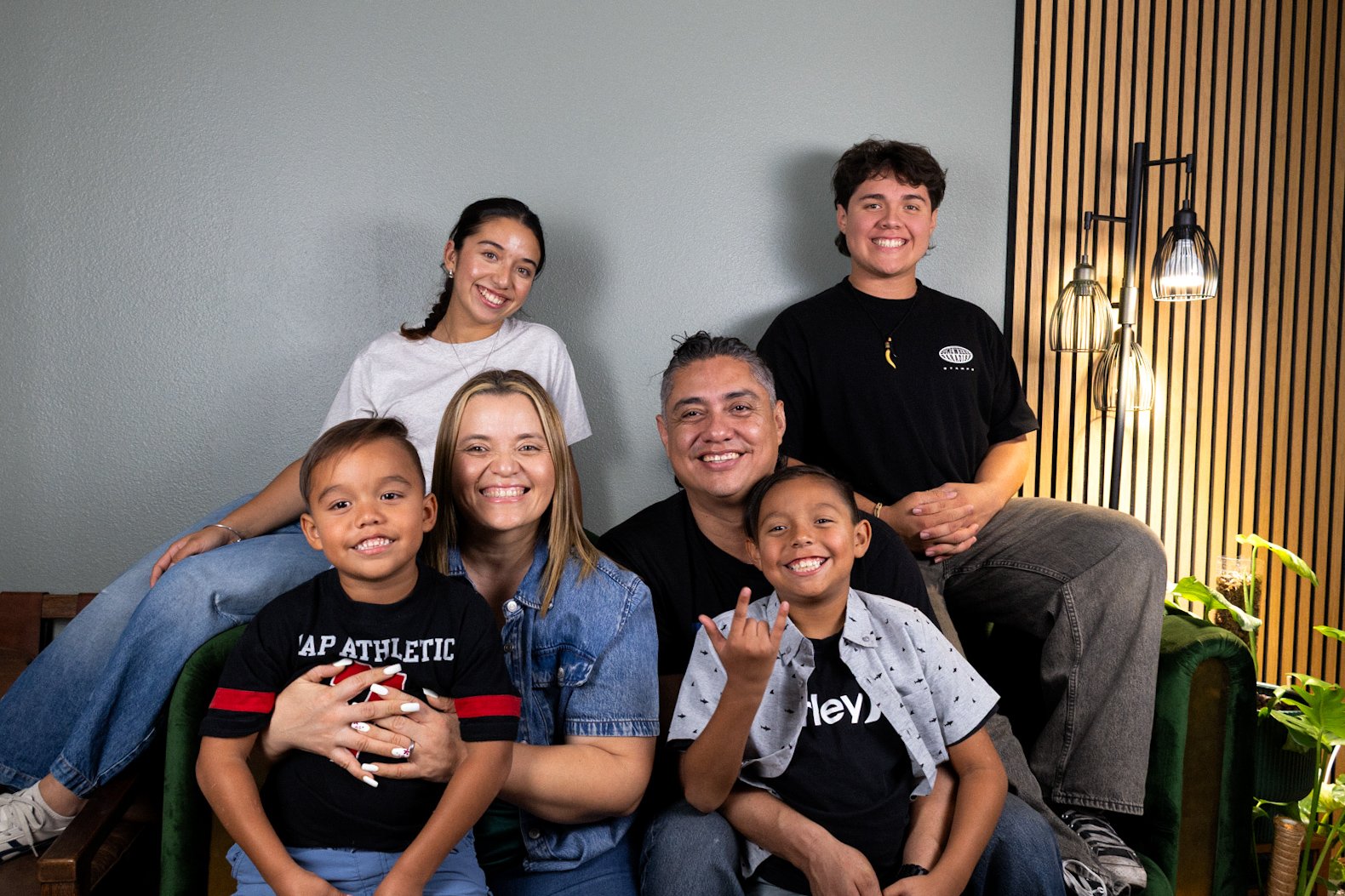 A multi-generational family of seven people smiling and posing together on a green couch in a cozy, modern living room with a gray wall, wooden panel accent wall, and stylish floor lamp.