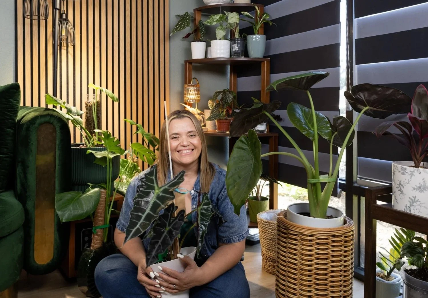 A woman sitting in a room filled with houseplants, smiling and holding a potted plant in a white pot, with a wooden and black accent wall in the background.