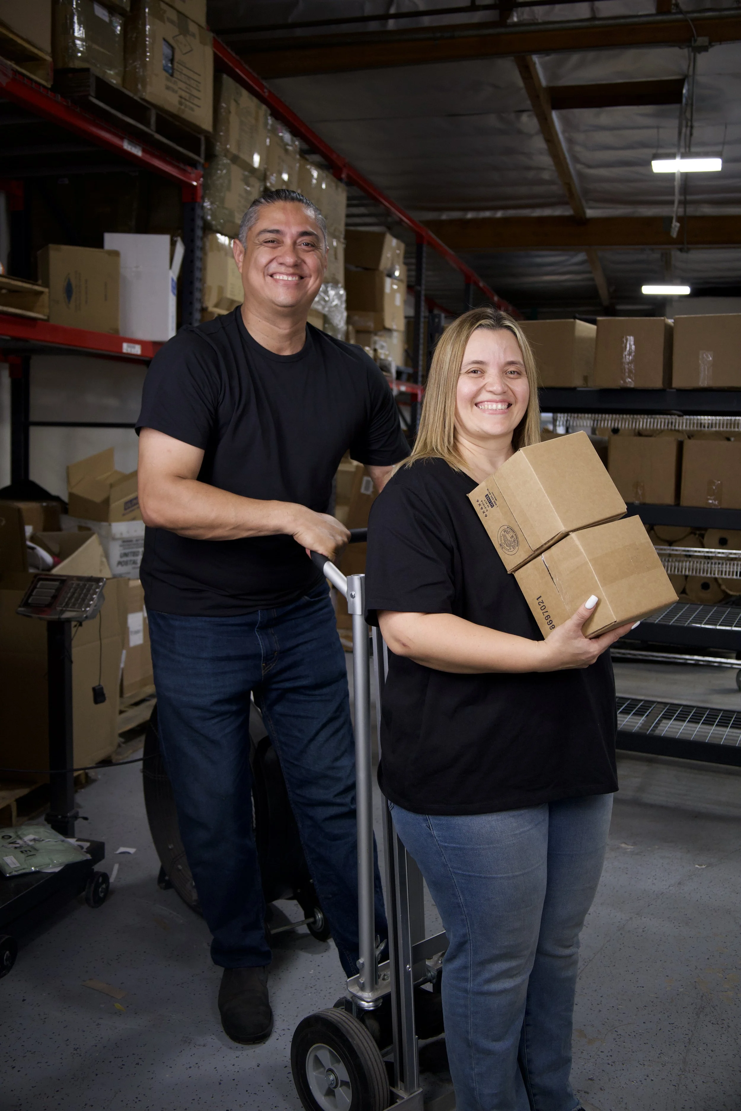 Two people stand in a warehouse surrounded by shelves filled with cardboard boxes. The woman in front is smiling and holding a small cardboard box, while the man behind her is smiling and pushing a hand truck. Both are wearing black shirts.