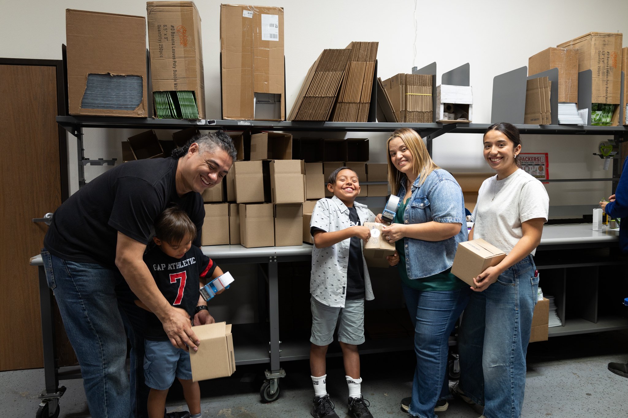A group of five people, including children and adults, are packing boxes in a room with shelves filled with cardboard boxes. They are smiling and working together.