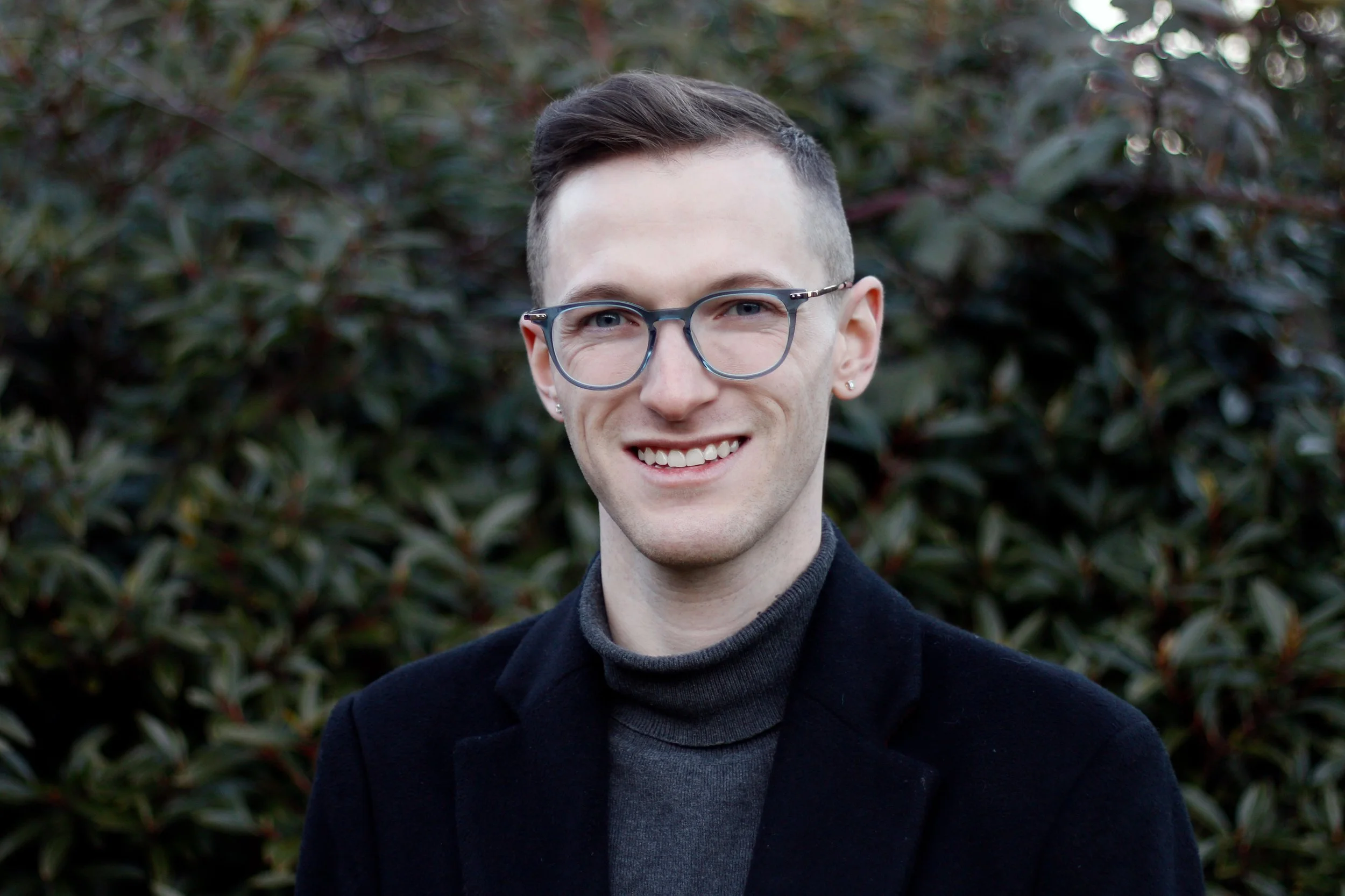 A young man with short brown hair, glasses, and earrings, smiling outdoors in front of green leafy bushes, wearing a dark jacket and turtleneck sweater.