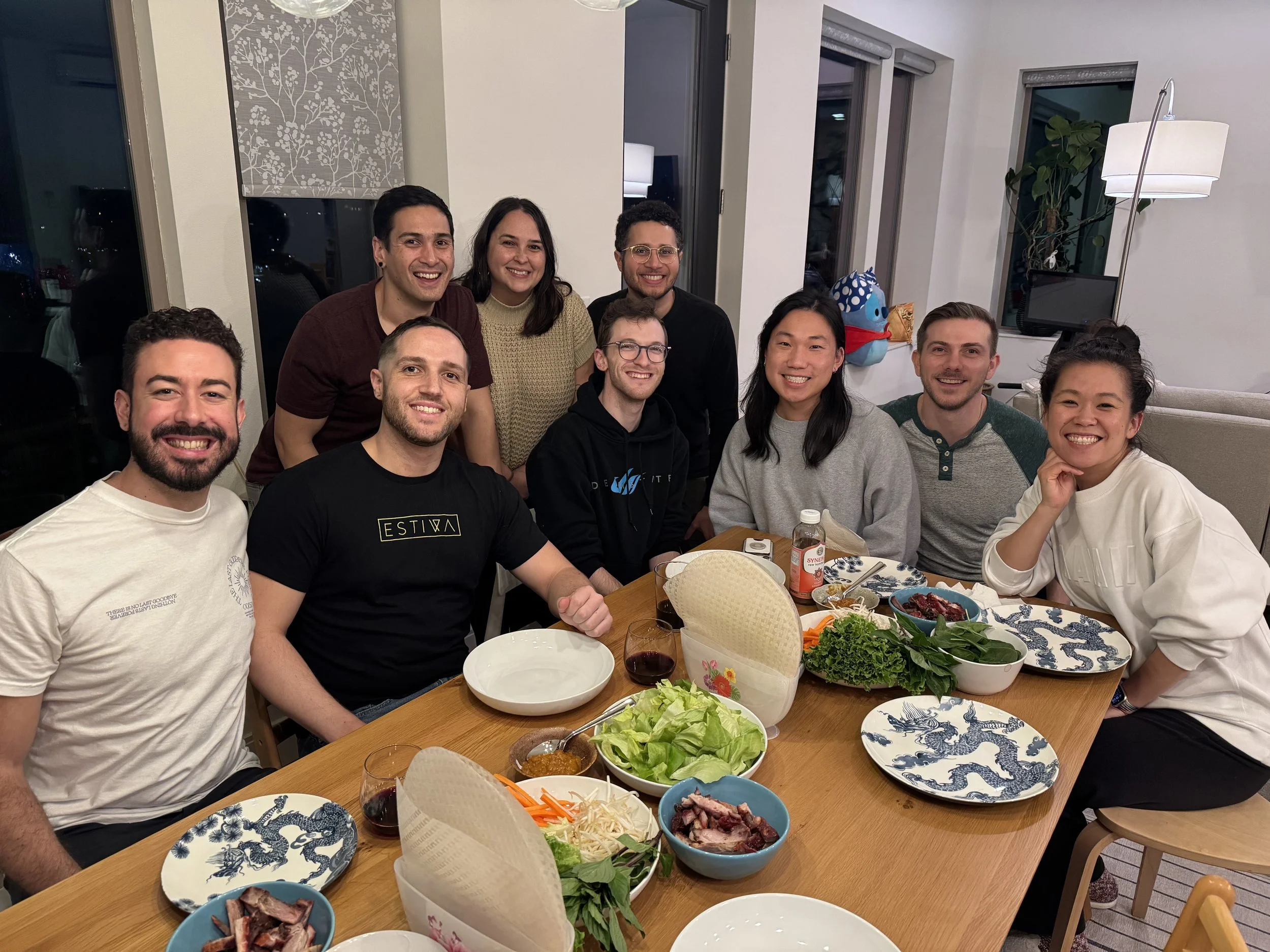 A group of nine people smiling around a dining table set with Asian style dishes, bowls of vegetables, meats, and sauces, in a modern living room with large windows and contemporary decor.