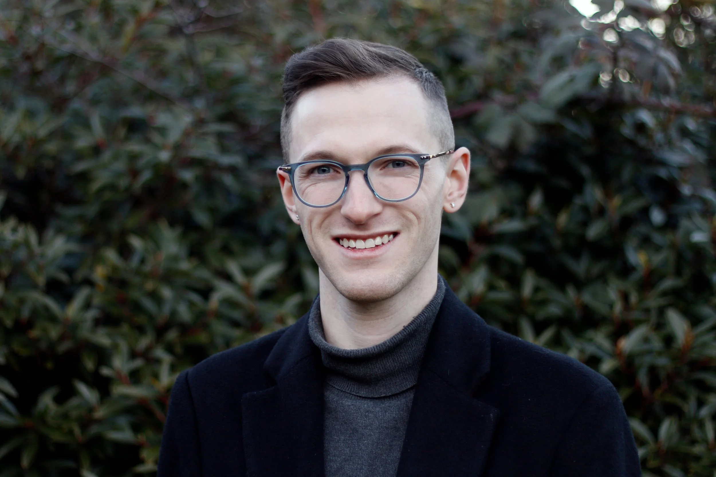 A young man with glasses smiling outdoors with greenery in the background.