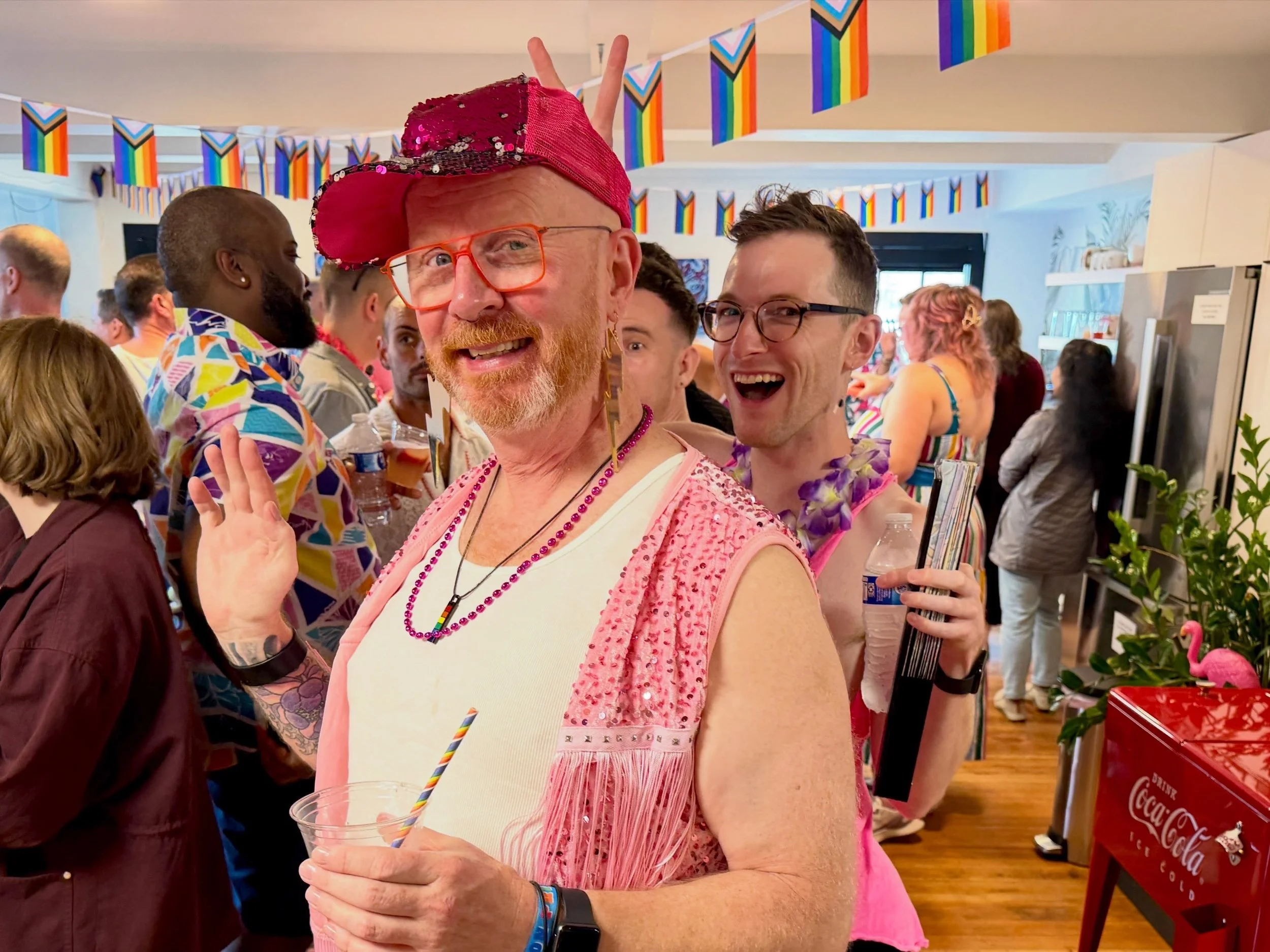 People celebrating at an LGBTQ+ pride event indoors, decorations include rainbow flags, with a man dressed in pink with rainbow beads, a sequined pink cap, and a pink vest, making a peace sign behind another smiling man with glasses.