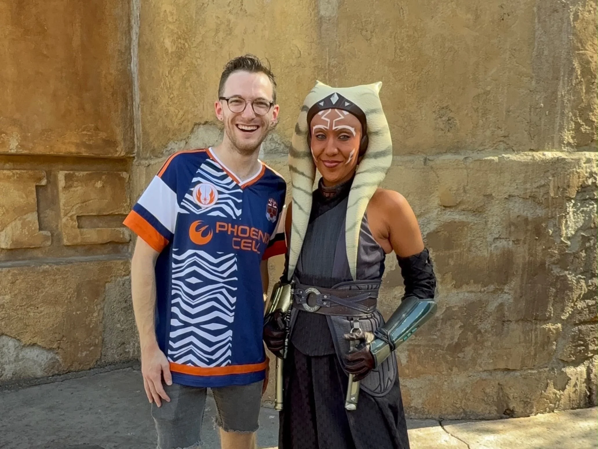 A young man wearing glasses and a blue sports jersey standing next to someone dressed as Ahsoka Tano from Star Wars, in front of a stone wall, smiling.