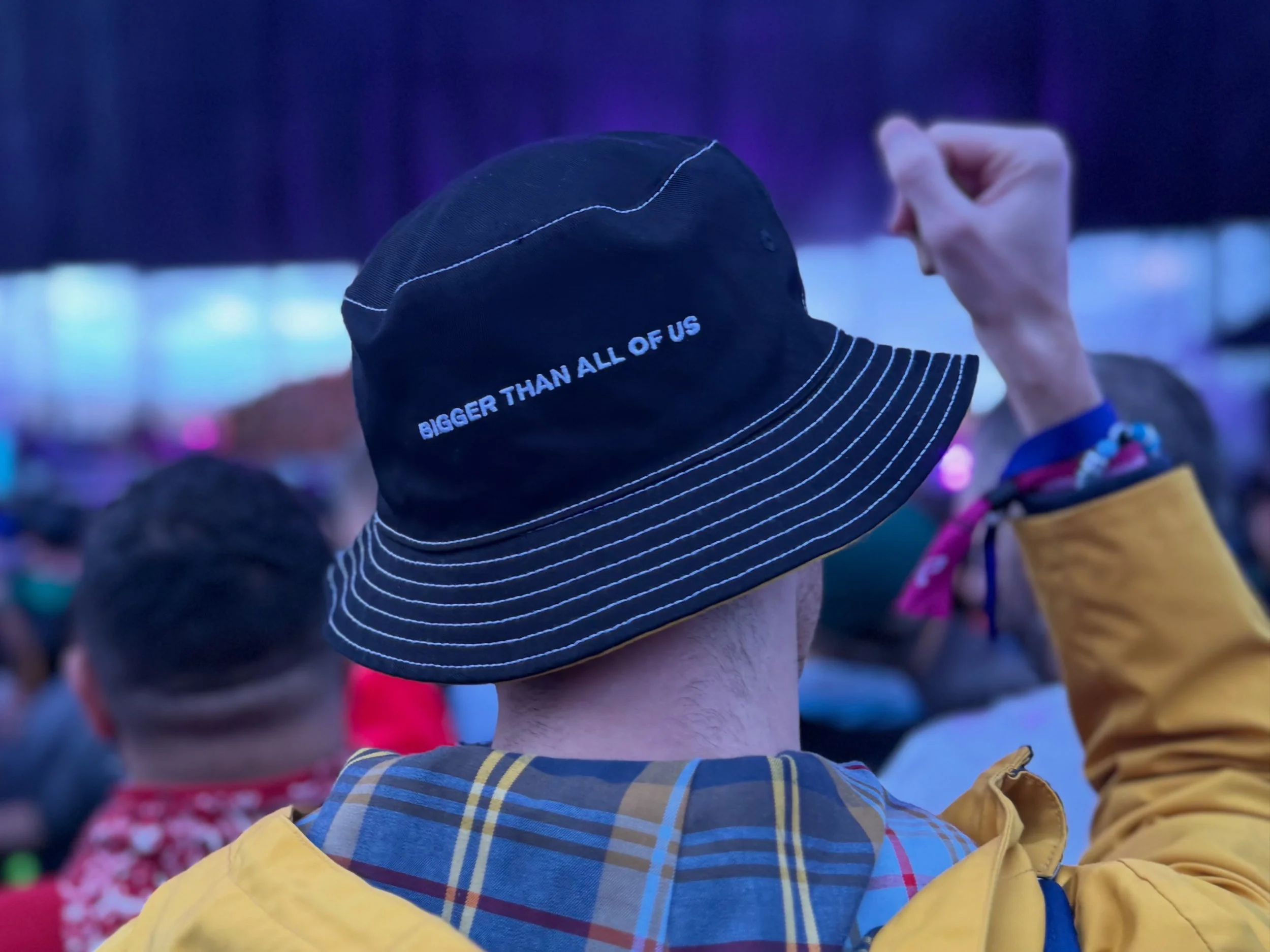 Close-up of a person wearing a black baseball cap with white stitching and the text "BIGGER THAN ALL OF US" on the side, reaching upward in a crowded indoor setting.
