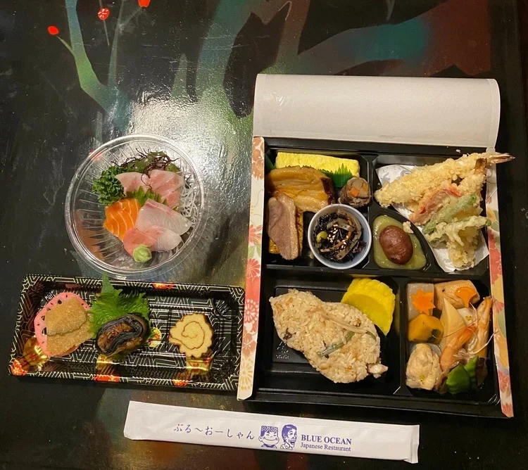 A Japanese meal with assorted sashimi, cooked vegetables, tempura, and other traditional dishes in a black compartmentalized tray, accompanied by a small plate of sashimi and condiments, and a pair of chopsticks with a paper sleeve from Blue Ocean Ja