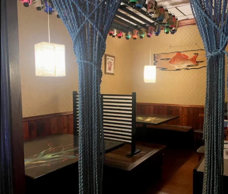 Interior of a Japanese-style restaurant with dark wood booths, hanging paper lanterns, and a blue rope curtain. Decorative elements include a fish artwork on the wall and colorful hanging decorations from the ceiling.