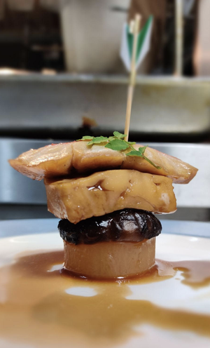 A gourmet appetizer featuring a skewer with slices of fish or meat topped with green herbs, set on a sauce with a dark mushroom or olive piece and a round base, garnished with additional herbs.