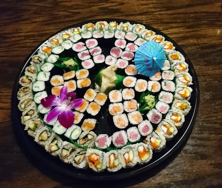 A round sushi platter with various types of sushi rolls, garnished with a purple orchid, a small paper umbrella, and green garnishes, placed on a wooden table.
